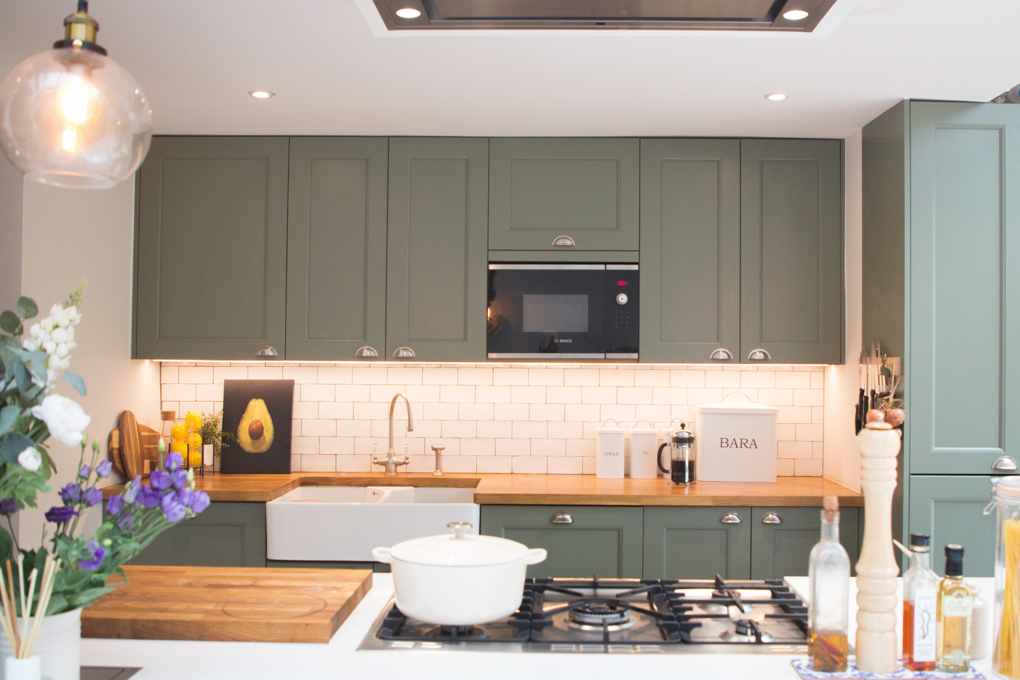 Modern kitchen featuring dark green cabinetry, a white farmhouse sink, and wooden countertops. The space includes a built-in microwave, stylish decor, and a gas stove, creating a functional and inviting cooking area. An Arch KBB team member is present, showcasing the design's practicality and aesthetic appeal.