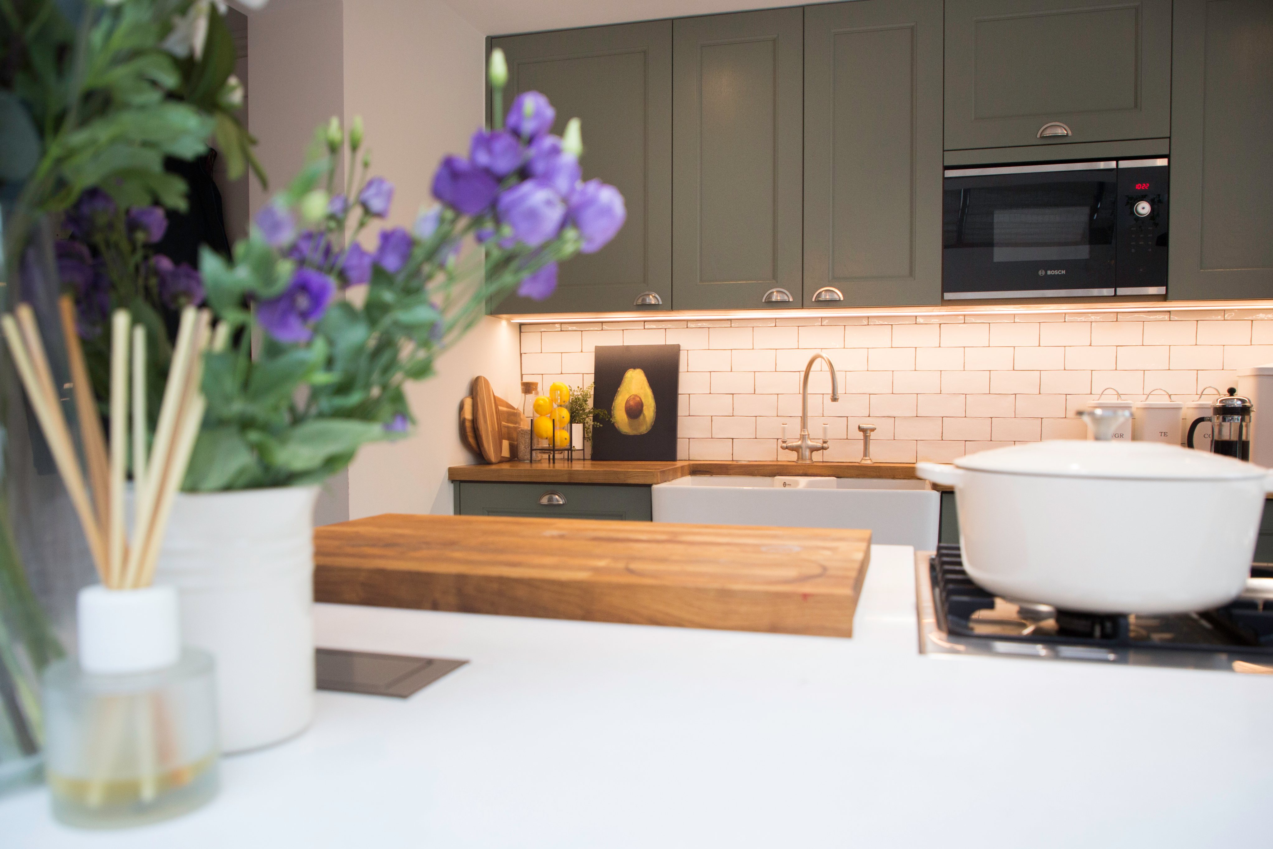 Modern kitchen interior featuring green cabinetry, a wooden countertop, and a minimalist design. A bouquet of purple flowers adds a decorative touch, while an Arch KBB team member is present in the background. The space is well-lit with a stylish backsplash and contemporary appliances.