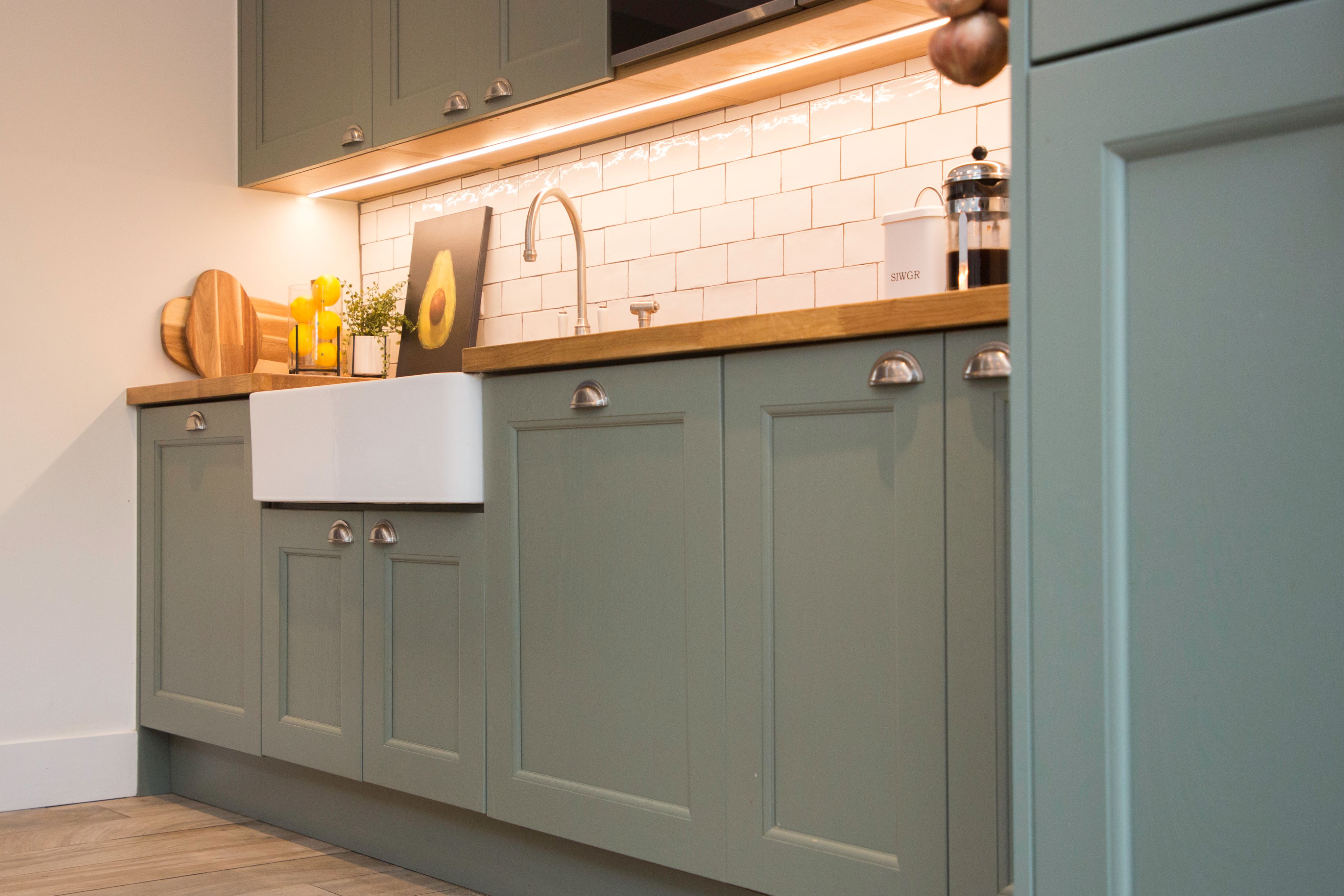 Modern kitchen featuring green cabinetry, a farmhouse sink, and a wooden countertop. The space is illuminated by under-cabinet lighting, highlighting a stylish backsplash of white tiles. Decorative items, including a cutting board and potted plant, add a touch of warmth. An Arch KBB team member is present, enhancing the overall design.