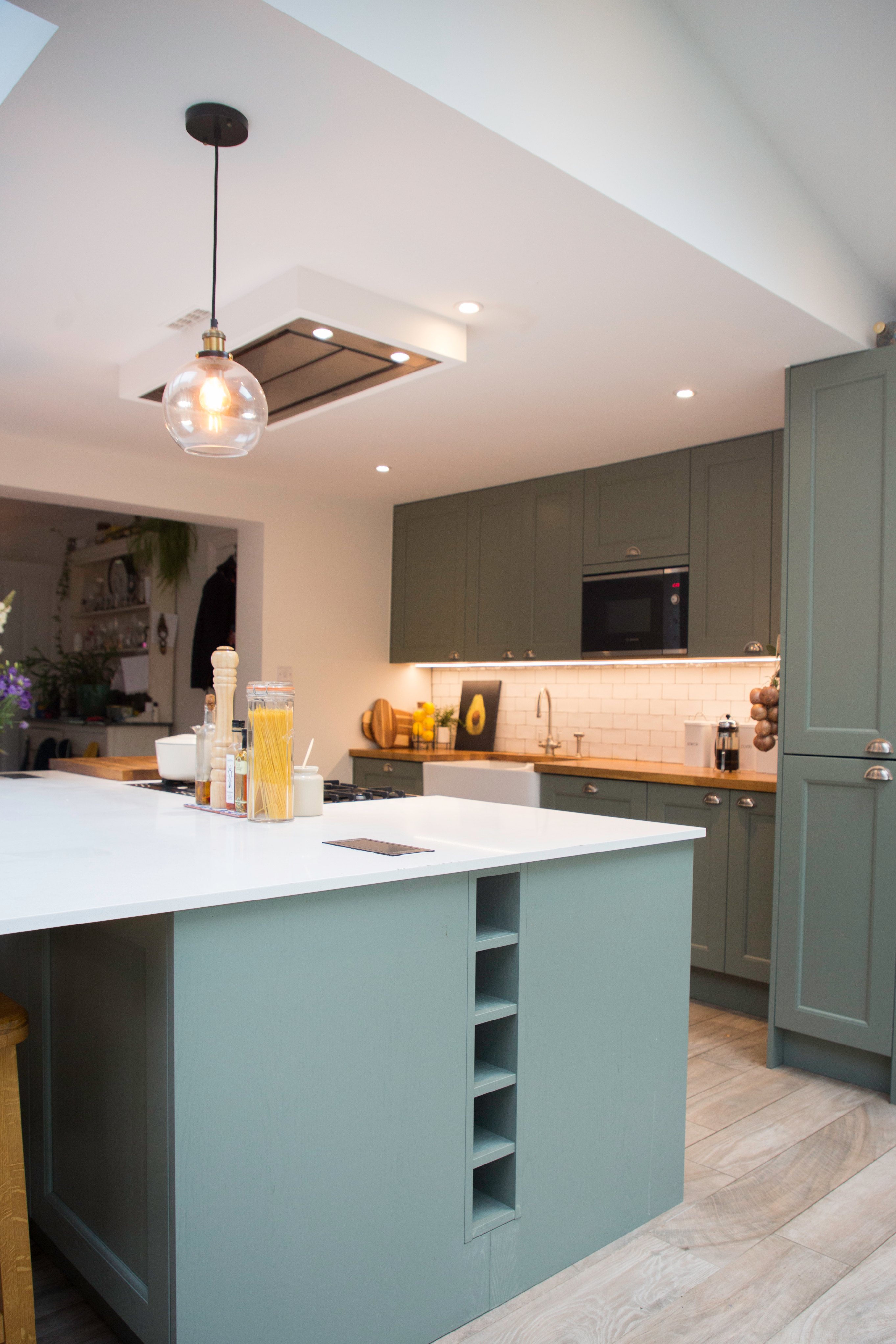 Modern kitchen design featuring a spacious island with a white countertop, green cabinetry, and wooden accents. The room is well-lit with pendant lighting and includes appliances such as a built-in oven and a stylish backsplash. An Arch KBB team member is present, showcasing the kitchen's functionality and aesthetic appeal.