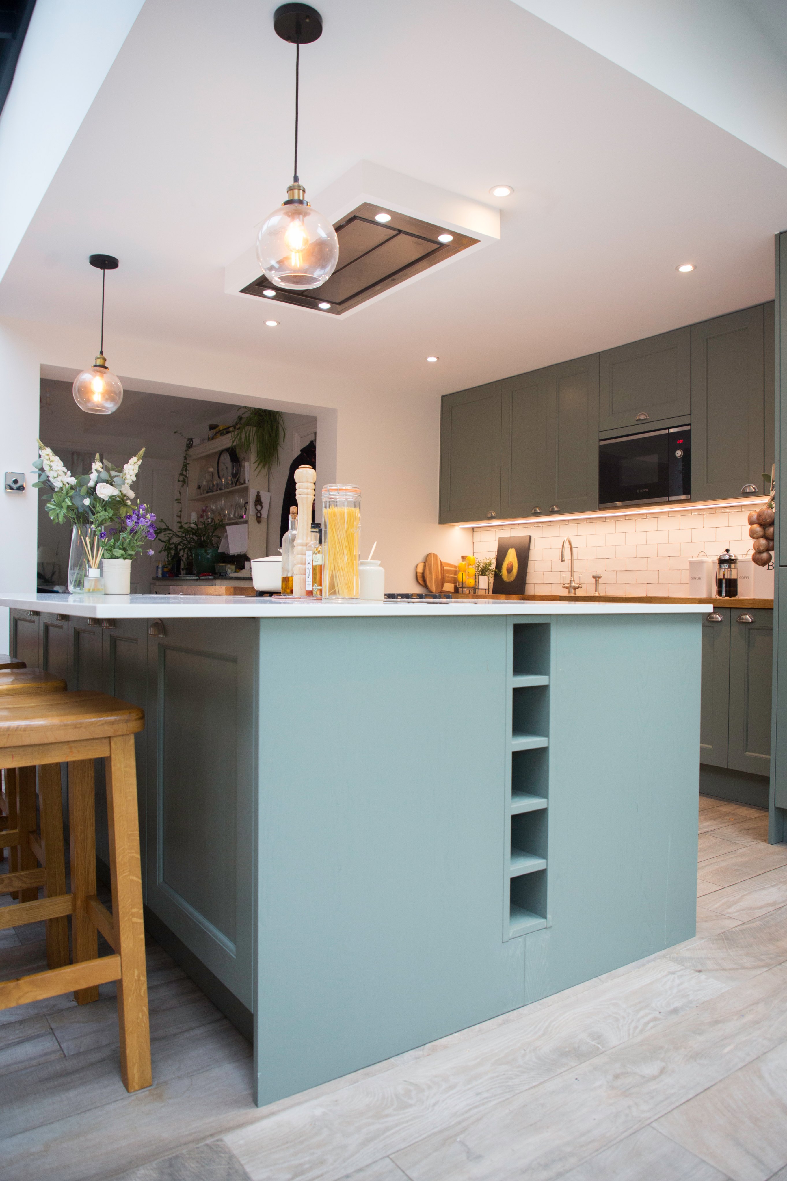 Modern kitchen design featuring a spacious island with a light blue finish, wooden bar stools, and stylish cabinetry. The countertop displays various kitchen items, including plants and bottles, while soft lighting enhances the inviting atmosphere. An Arch KBB team member is engaged in the space, showcasing a contemporary and functional kitchen environment.