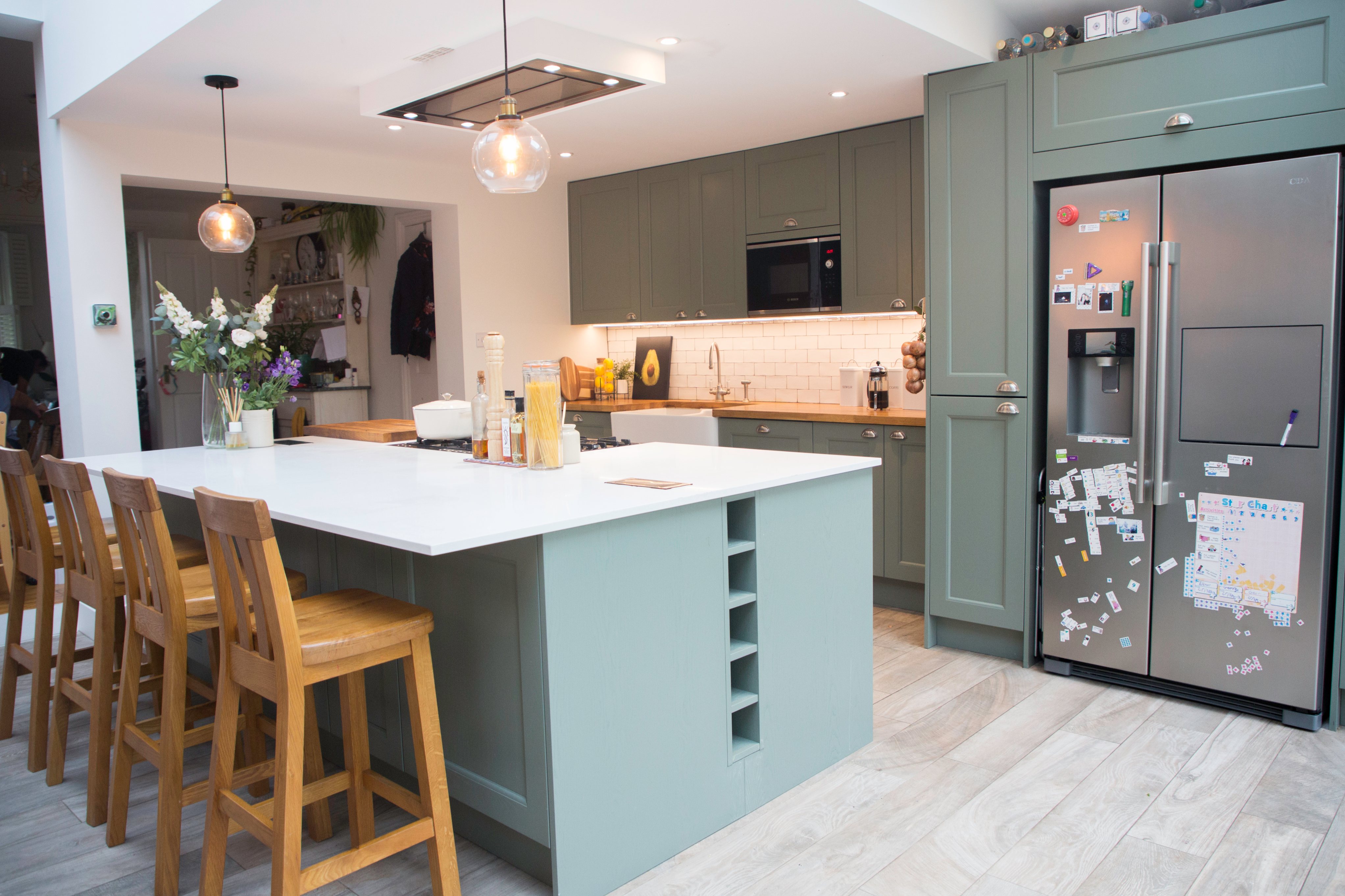 Modern kitchen featuring a large island with seating, light green cabinetry, and stainless steel appliances. An Arch KBB team member is present, showcasing a stylish and functional design. The space includes decorative lighting and a mix of natural and artificial elements, creating an inviting atmosphere.