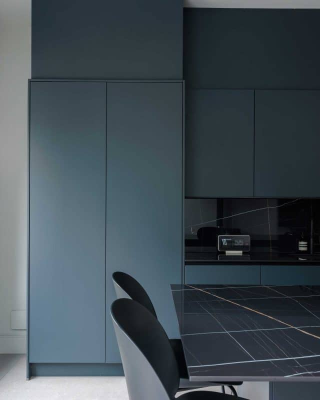 Modern kitchen featuring sleek blue cabinetry and a stylish black countertop. An Arch KBB team member is present, providing design insights. The space showcases contemporary design elements and a minimalist aesthetic.