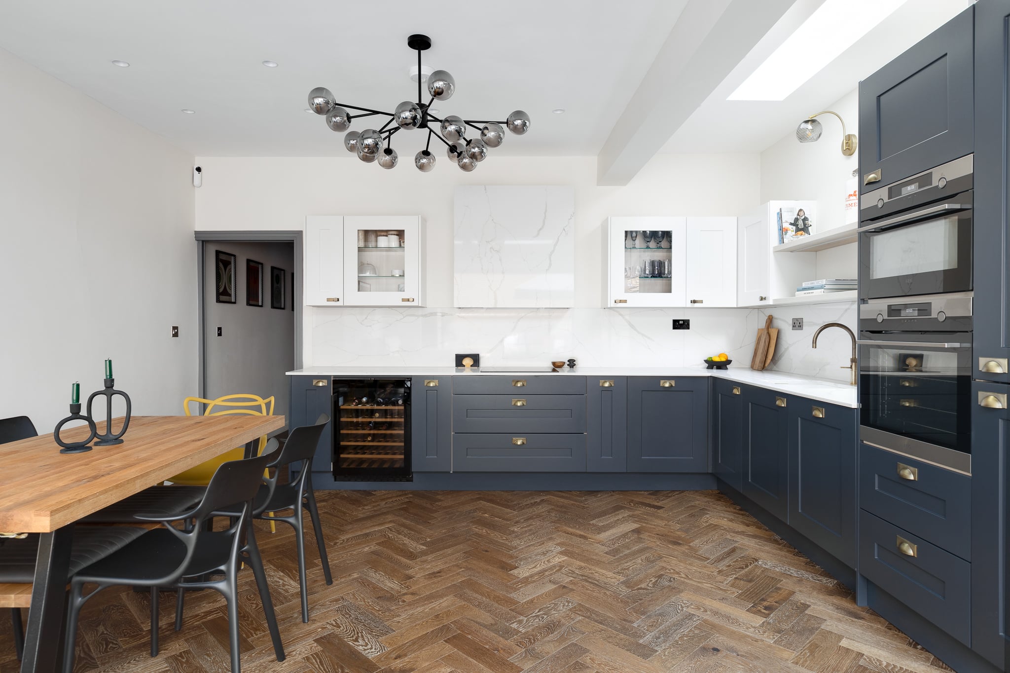 Modern kitchen design featuring a stylish combination of navy blue and white cabinetry, a wooden dining table with colourful chairs, and herringbone patterned flooring. The space is illuminated by a contemporary chandelier and includes built-in appliances, creating a functional and aesthetically pleasing environment. An Arch KBB team member is present, showcasing the kitchen's layout.