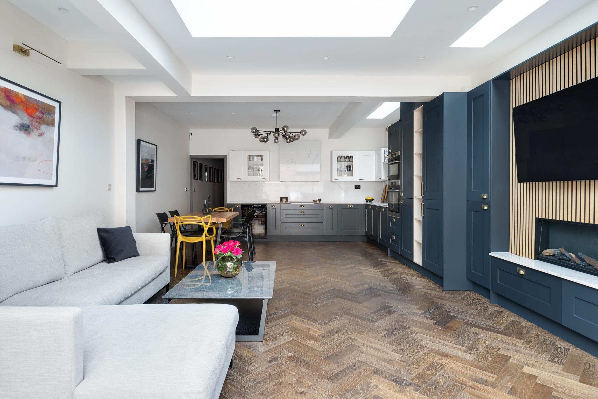 Modern interior design featuring an open-plan living space with a stylish sofa, a coffee table, and a kitchen with blue cabinetry. Natural light floods the room through a skylight, highlighting the herringbone wooden flooring. An Arch KBB team member is present, contributing to the vibrant atmosphere enhanced by decorative plants and contemporary furnishings.