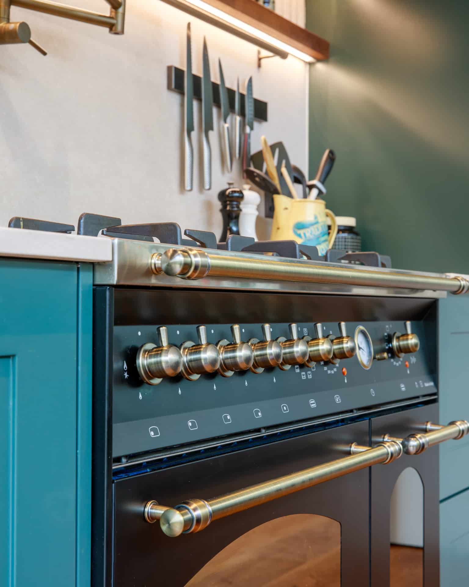 Close-up of a stylish gas range with brass knobs and a pressure gauge, set against a modern kitchen backdrop featuring neatly arranged utensils and cookware, showcasing the sophisticated design of Arch KBB kitchen installations.