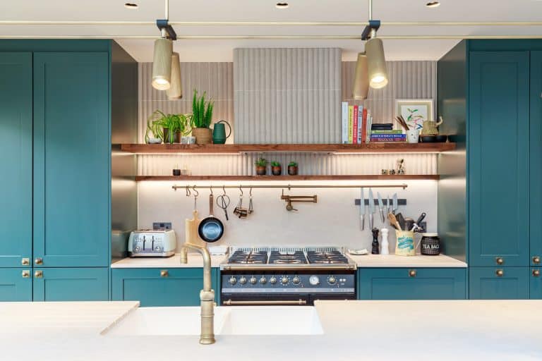 Modern kitchen design featuring teal cabinetry, a stylish gas range, and open shelving displaying kitchenware and plants. The space is well-lit with pendant lights and has a sleek white countertop. An Arch KBB team member is present, showcasing the contemporary kitchen layout and design elements.