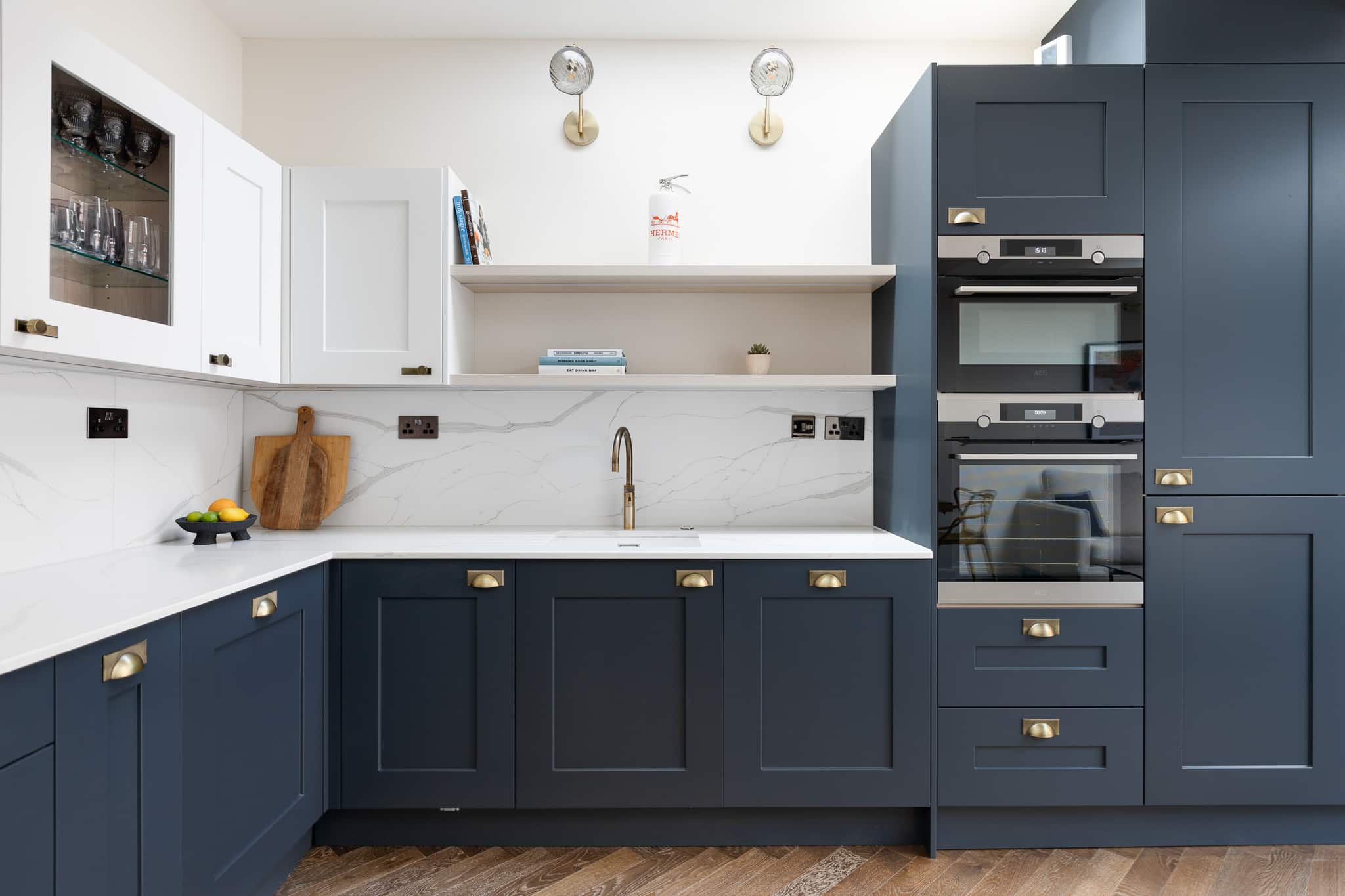 Modern kitchen design featuring navy blue cabinetry, a marble splashback, and integrated appliances. The layout includes a sleek sink and wooden flooring, creating a stylish and functional space. An Arch KBB team member is present, showcasing the kitchen's features.