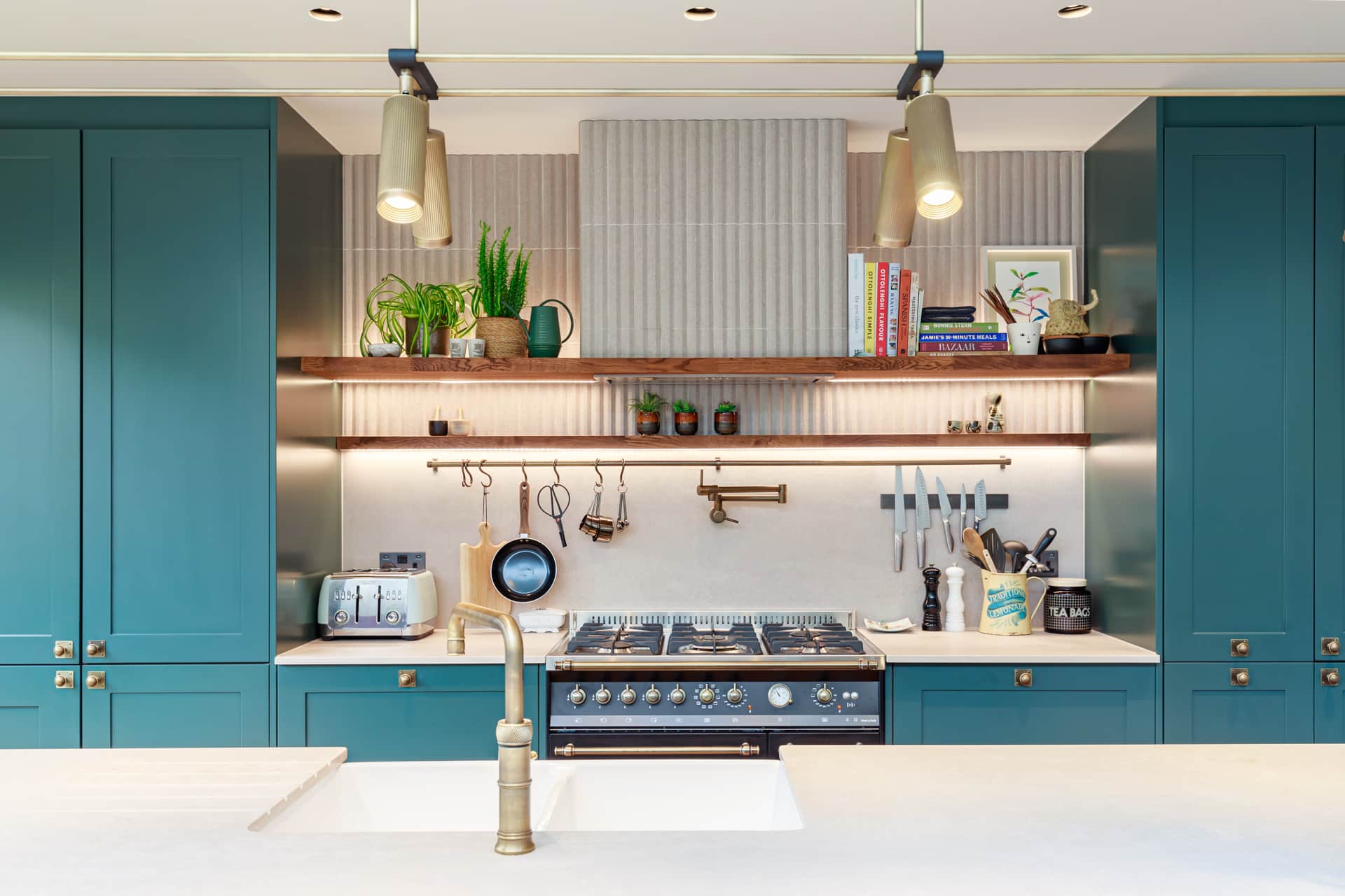 Modern kitchen design featuring teal cabinetry, a stylish gas range, and open shelving displaying kitchenware and plants. The space is well-lit with pendant lights and has a sleek white countertop. An Arch KBB team member is present, showcasing the contemporary kitchen layout and design elements.