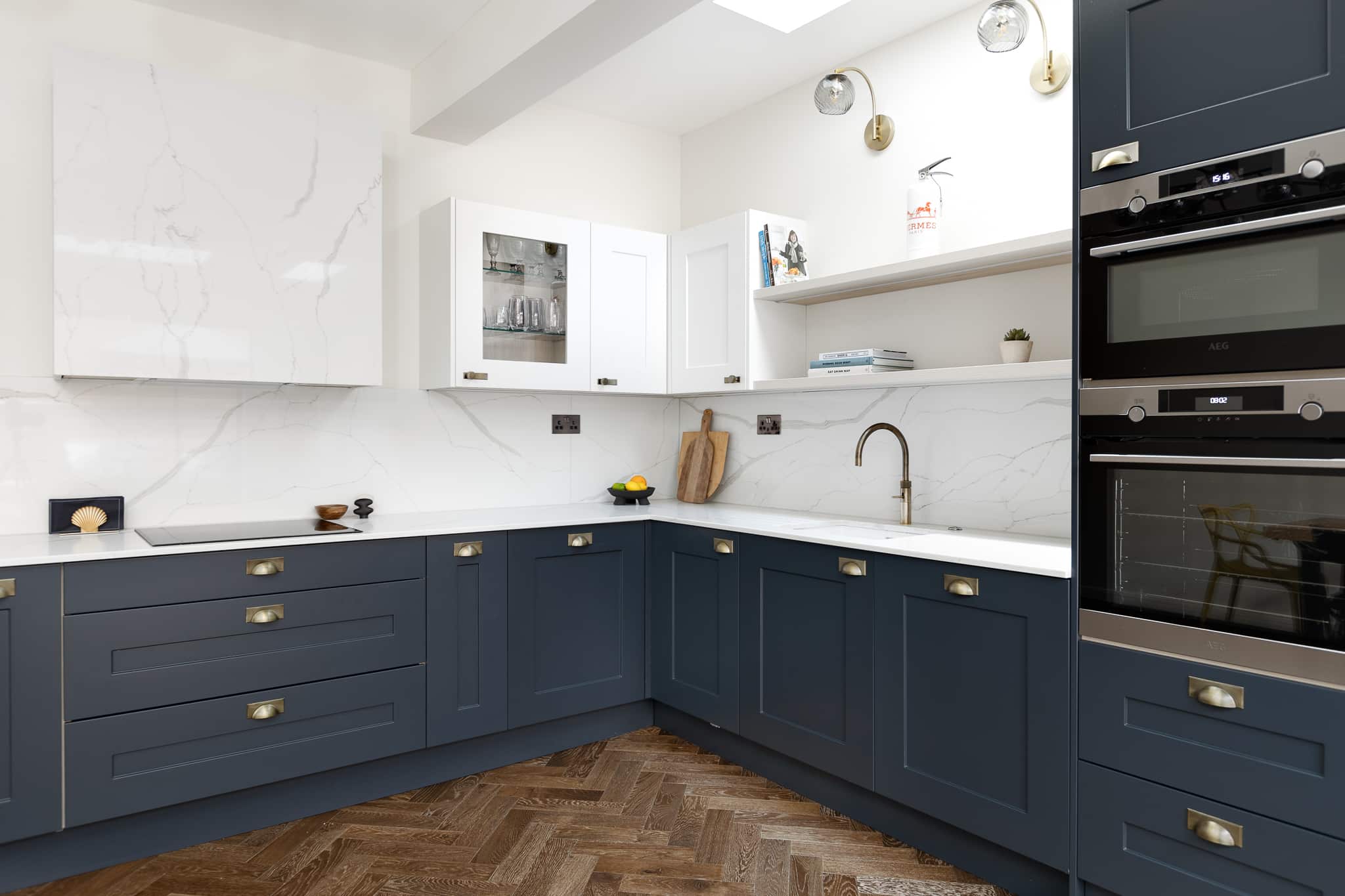 Modern kitchen design featuring navy blue and white cabinetry with marble countertops, stylish brass handles, and built-in appliances. The space includes a practical layout with ample storage, a sleek sink, and a wooden herringbone floor. An Arch KBB team member is present, showcasing the kitchen's functionality and aesthetic appeal.