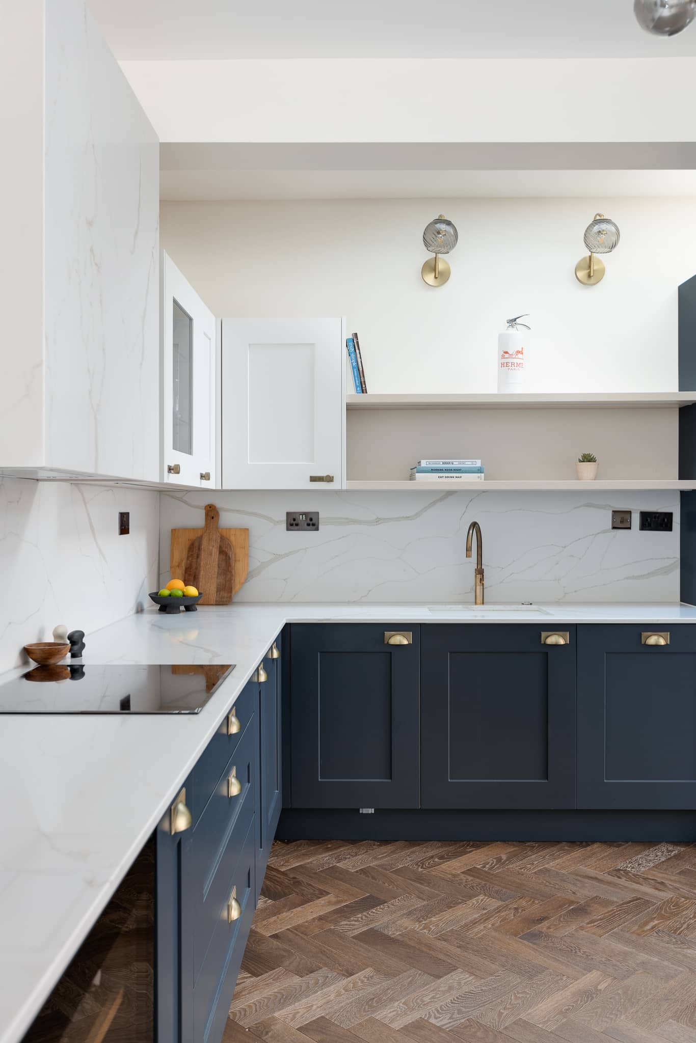 Modern kitchen design featuring a sleek white and navy colour scheme. The image showcases a stylish countertop with a built-in hob, elegant cabinetry, and brass fixtures. A minimalist shelf displays books and a small plant, enhancing the contemporary aesthetic. An Arch KBB team member can be seen, contributing to the kitchen's design.
