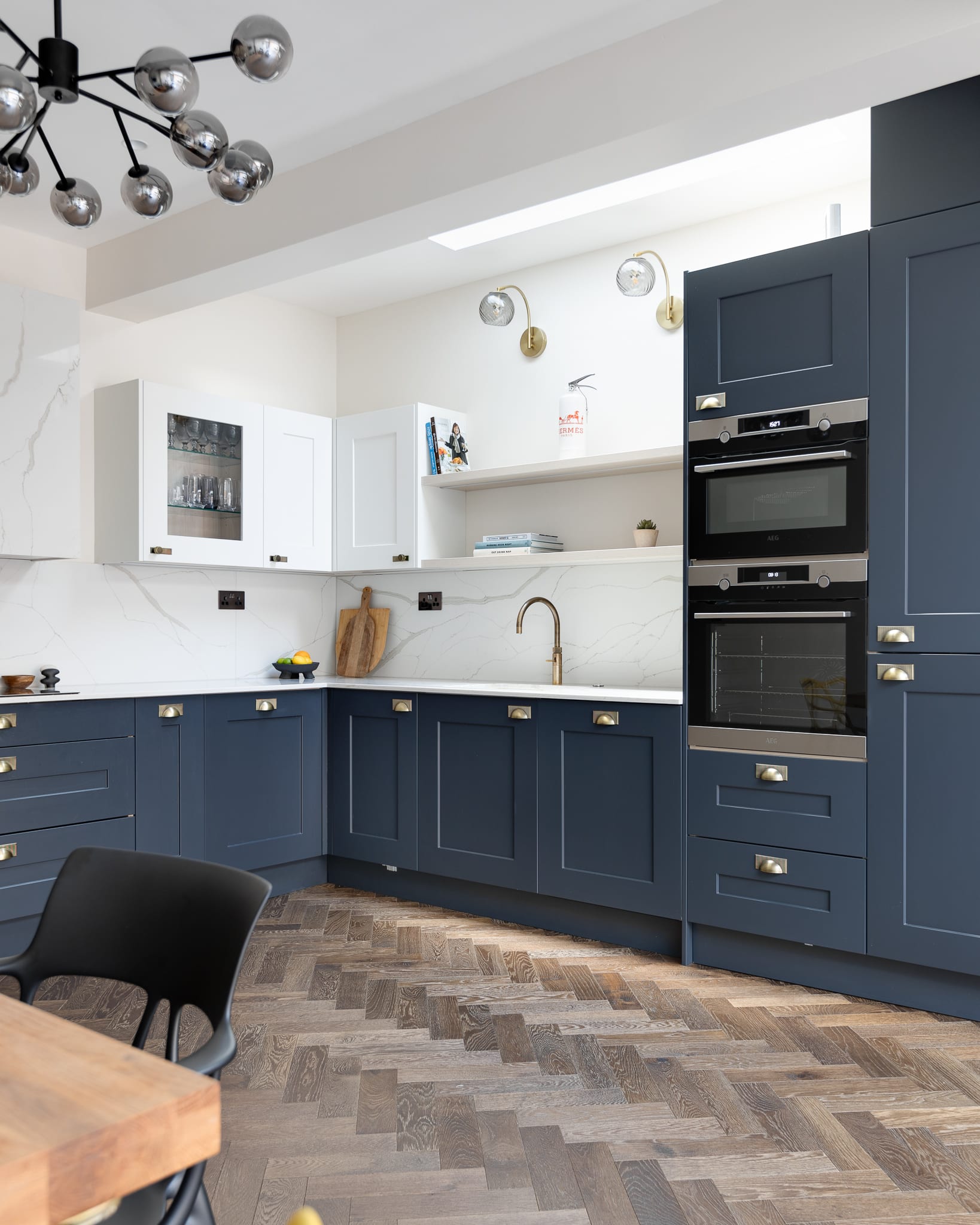 Modern kitchen design featuring a stylish combination of navy blue and white cabinetry, marble backsplash, and integrated appliances. The space includes a wooden chopping board, decorative items on the shelves, and elegant brass fixtures, showcasing a contemporary aesthetic. An Arch KBB team member is present, enhancing the kitchen's inviting atmosphere.