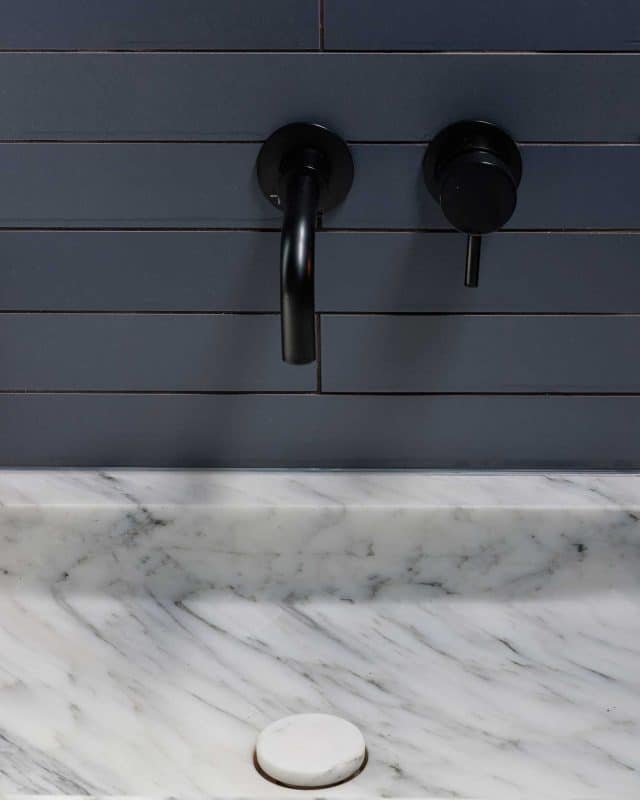 Close-up of a stylish bathroom sink featuring a sleek marble countertop and modern black taps against a dark grey tiled wall, showcasing contemporary design elements. An Arch KBB team member is present, contributing to the design process.