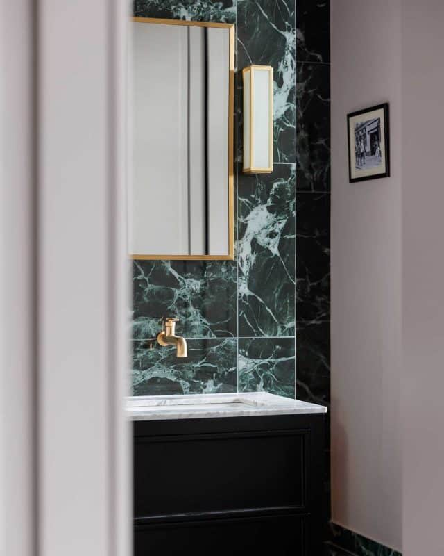 A stylish bathroom featuring dark green marble tiles, a sleek gold tap, and a modern mirror. The elegant design is complemented by a marble countertop and framed artwork on the wall, creating a sophisticated atmosphere. An Arch KBB team member is visible, enhancing the scene with their presence.