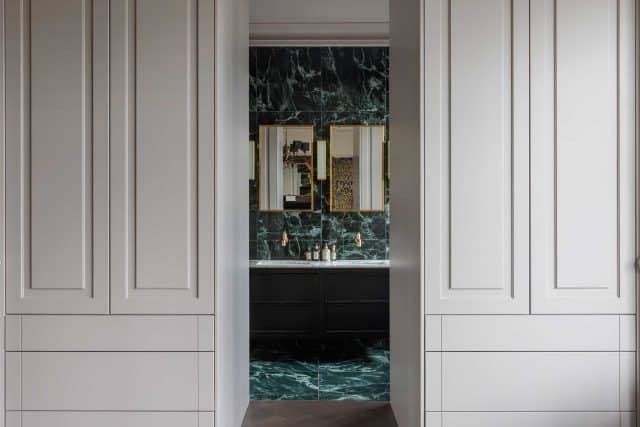 Interior view of a stylish bathroom featuring dark cabinetry and a striking green marble wall, framed by elegant mirrors and gold fixtures, with an Arch KBB team member visible.