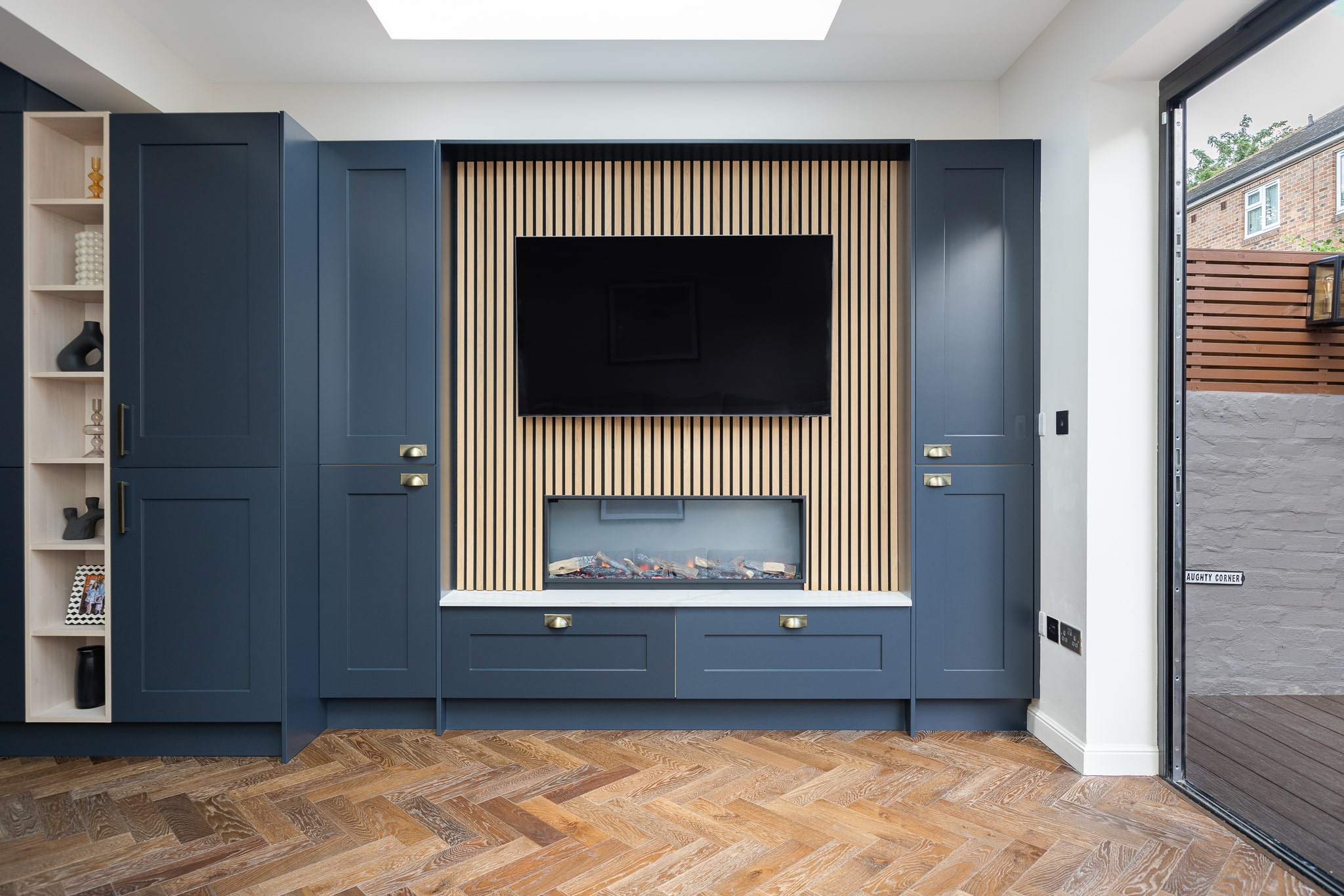 Modern living room featuring a stylish media unit with a wall-mounted television and an integrated electric fireplace. The unit is flanked by deep blue cabinetry and accented with wooden slats, complemented by a herringbone wooden floor. An Arch KBB team member is present, showcasing the elegant design.