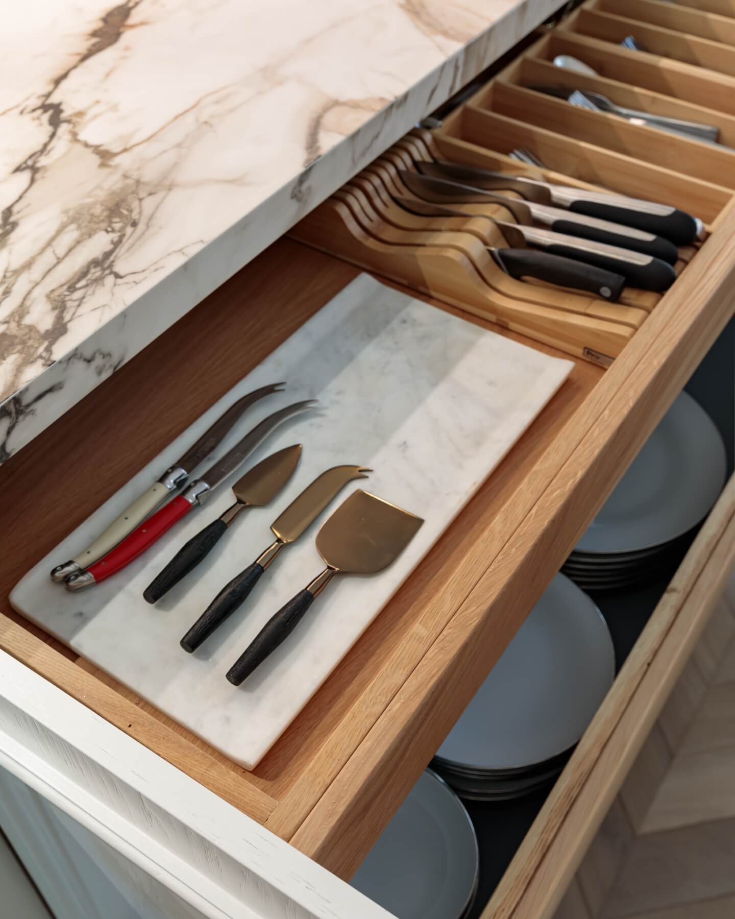 Interior view of a wooden kitchen drawer featuring a marble serving board and an assortment of cheese knives, including a curved knife, a cheese plane, and other cutting tools, alongside neatly arranged plates. An Arch KBB team member is present, contributing to the kitchen's organised aesthetic.