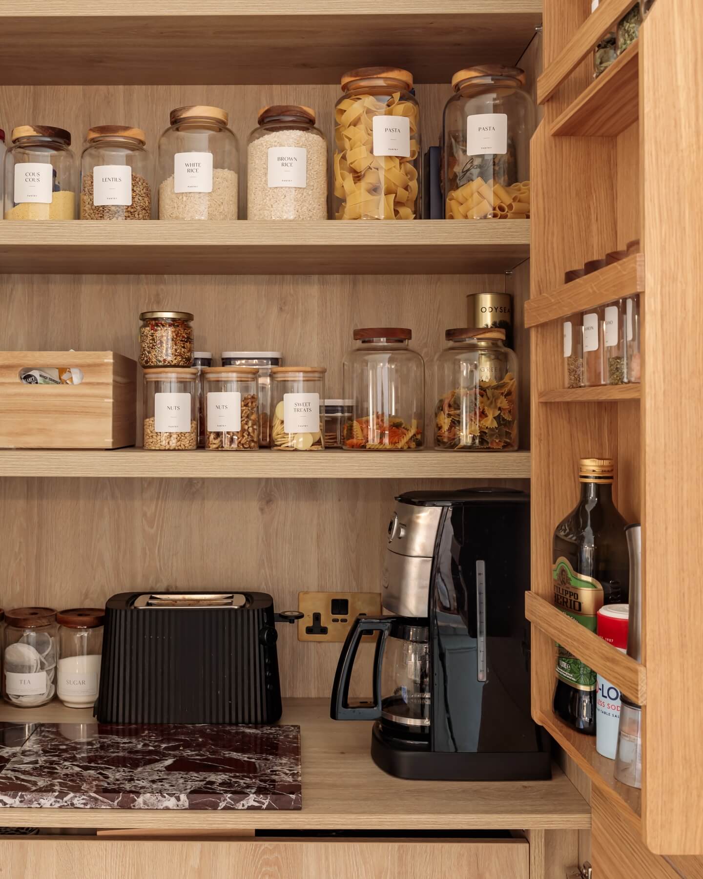 A well-organised kitchen pantry featuring glass jars with labelled contents, including grains, pasta, and dried herbs. An Arch KBB team member is present, alongside a toaster, coffee maker, and olive oil on the shelves, showcasing a functional and stylish storage solution.