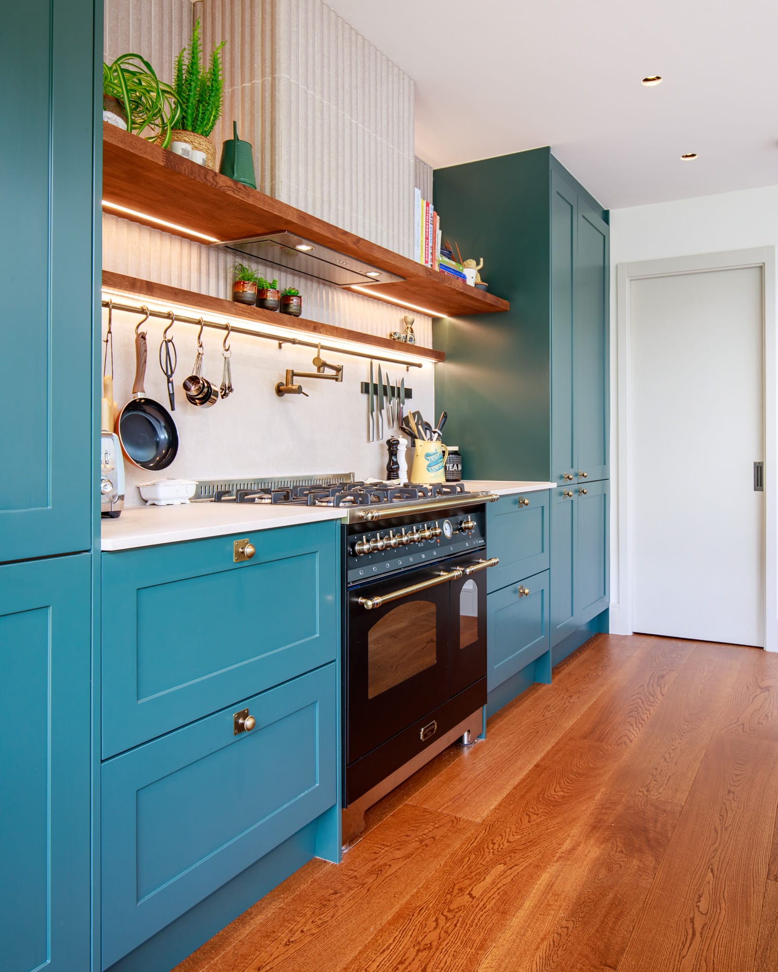 Modern kitchen design featuring teal cabinetry, a black oven, and stylish brass fixtures. The space includes a gas hob, hanging utensils, and decorative shelves with potted plants, creating an inviting and functional cooking area. An Arch KBB team member is present, enhancing the kitchen's aesthetic appeal.