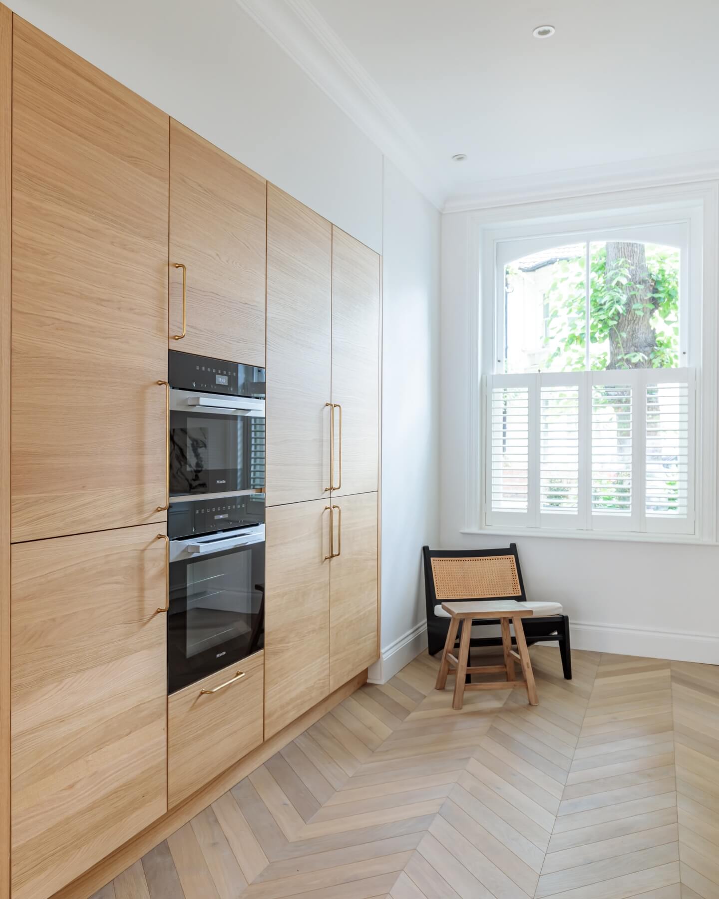 Modern kitchen featuring sleek wooden cabinetry with integrated black appliances, including double ovens. Natural light streams through a large window, illuminating a stylish seating area with a minimalist chair. An Arch KBB team member is present, enhancing the design atmosphere.
