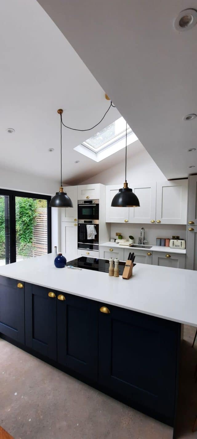 Modern kitchen design featuring a spacious layout with a central island, sleek cabinetry in muted tones, and stylish pendant lighting. The room is illuminated by natural light from a skylight and opens to a garden view through large glass doors. An Arch KBB team member is present, showcasing the kitchen's functionality and aesthetic appeal.