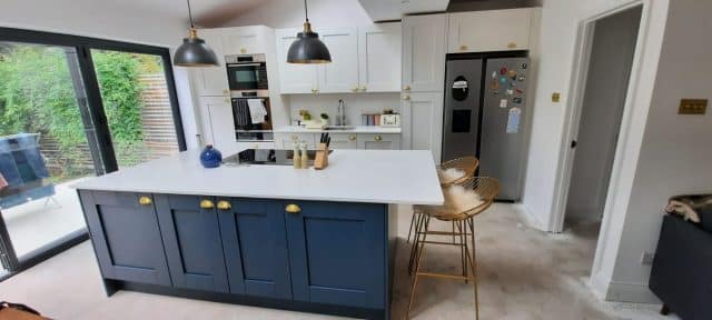 Modern kitchen design featuring a spacious island with a white countertop, blue cabinetry, and brass handles. The kitchen includes built-in appliances, stylish pendant lights, and a seating area with metallic bar stools. An Arch KBB team member is present, showcasing the inviting atmosphere of the space.