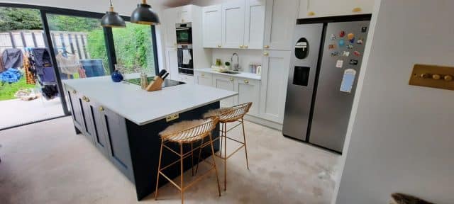 Modern kitchen featuring a large island with a white countertop, stylish bar stools, and sleek cabinetry. The space includes built-in appliances, a stainless steel fridge, and a window providing natural light, creating an inviting atmosphere. An Arch KBB team member is present, contributing to the design process.