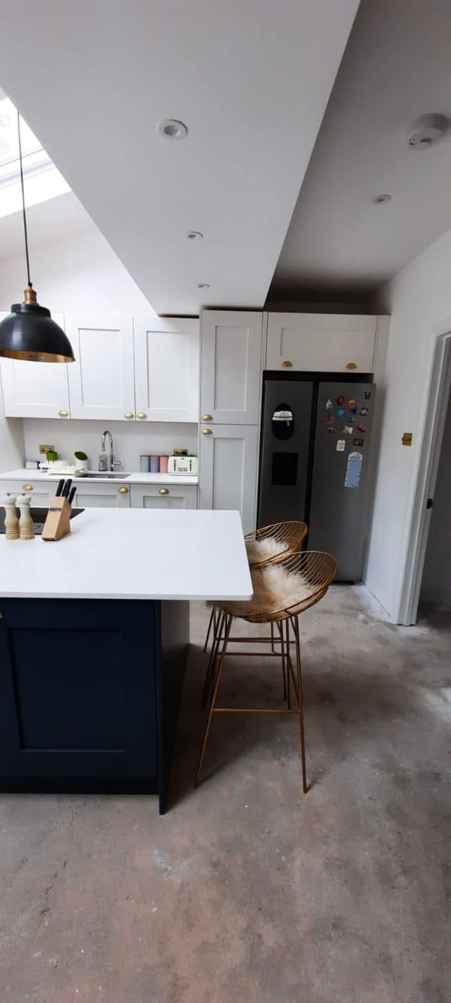 Modern kitchen featuring white cabinetry with brass handles, a sleek island with a white countertop, and stylish gold bar stools. Appliances include a stainless steel refrigerator adorned with magnets and a retro-style toaster. The space is well-lit and designed for functionality and aesthetic appeal, showcasing a blend of contemporary and minimalist elements. An Arch KBB team member is present, adding to the inviting atmosphere.