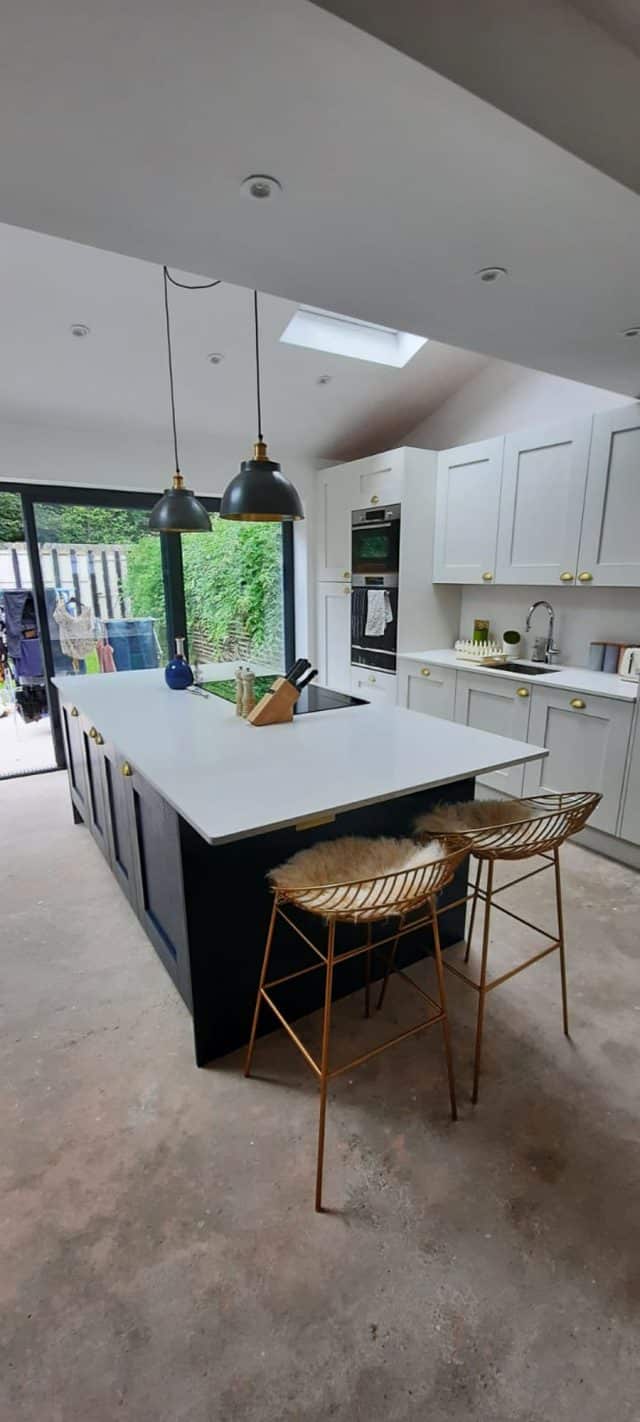 Modern kitchen featuring a spacious island with a white countertop, surrounded by stylish bar stools. The design includes grey cabinetry with gold handles, built-in appliances, and large windows that provide natural light and a view of the garden. An Arch KBB team member is present, showcasing the kitchen's functionality and aesthetic appeal.