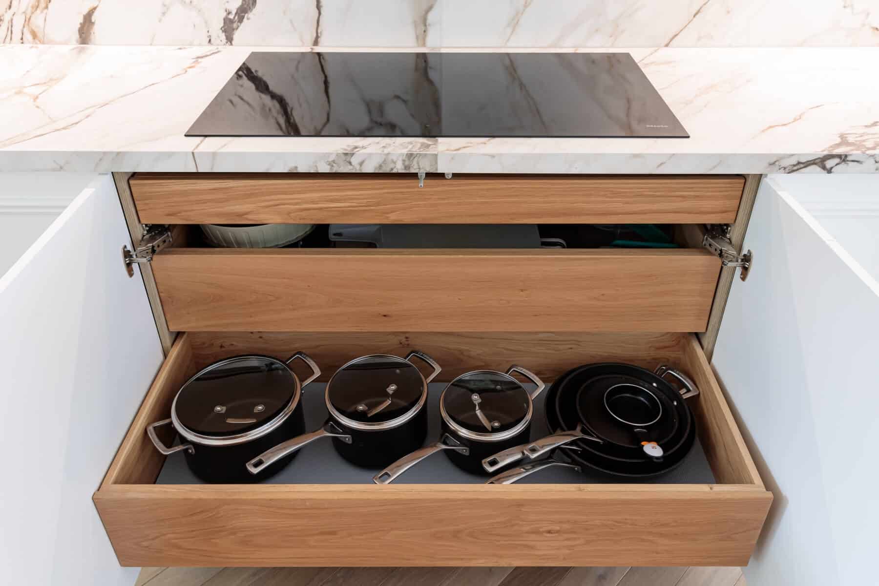 A well-organised kitchen drawer featuring black cookware, including pots and pans with lids, alongside plates. The drawer is made of light wood, complementing the marble countertop above. An Arch KBB team member is present, showcasing modern kitchen design and storage solutions.