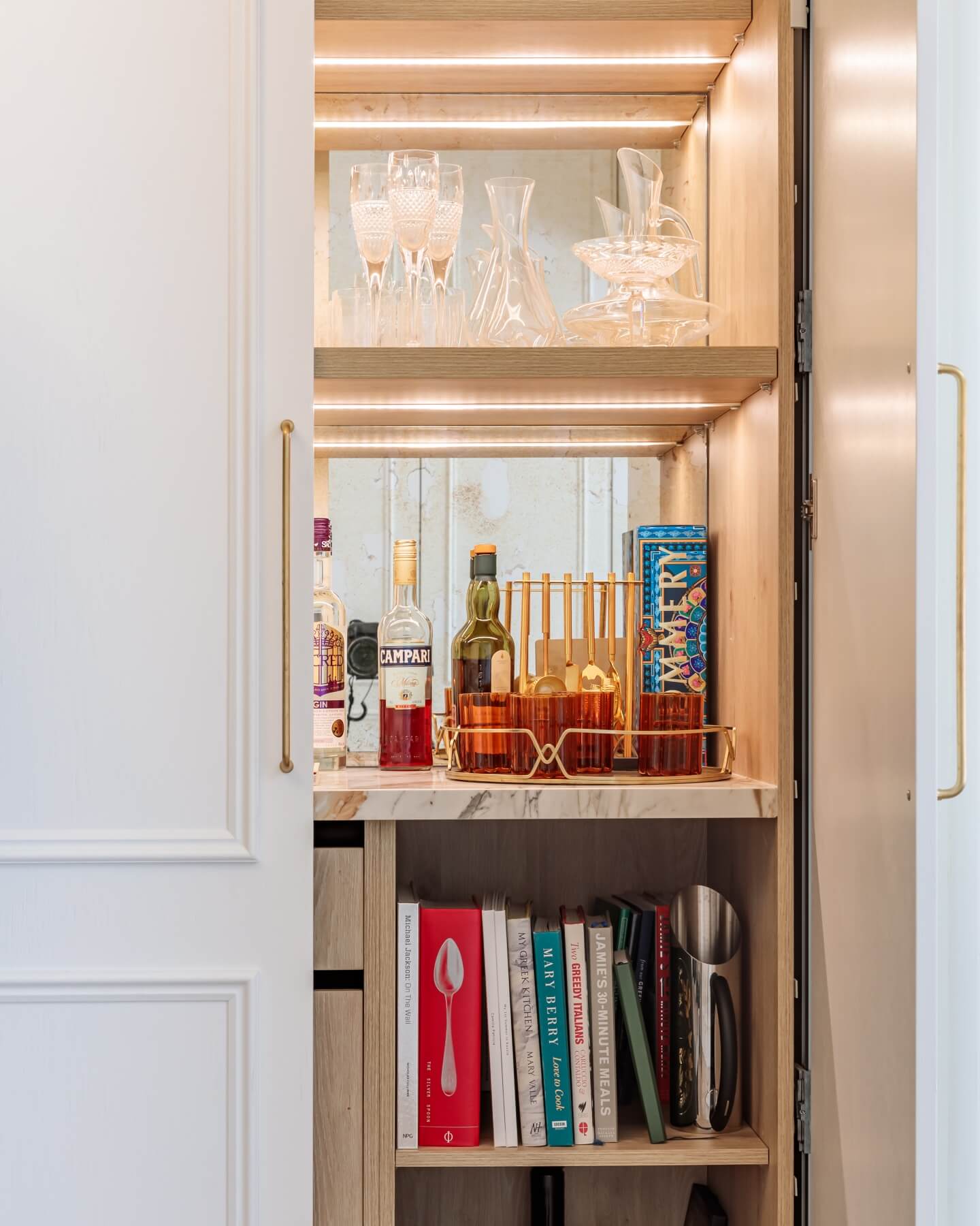 A stylish home bar cabinet featuring an array of spirits, elegant glassware, and cocktail accessories, with warm lighting highlighting the contents. An Arch KBB team member is present, showcasing the design.