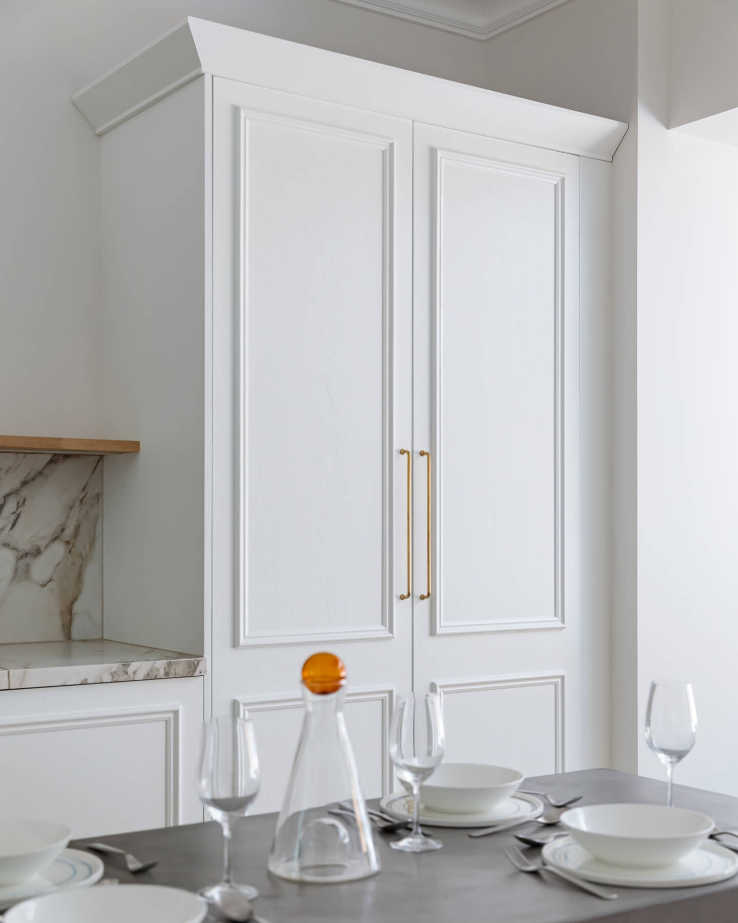 Modern kitchen featuring elegant white cabinetry with gold handles and a marble countertop, complemented by glassware and a decanter. An Arch KBB team member is present, showcasing the stylish interior design.