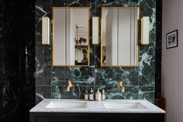 Arch KBB team member standing in a stylish bathroom featuring a double sink with a marble countertop, surrounded by striking green marble walls. The space is illuminated by elegant wall sconces and showcases two rectangular mirrors with gold frames, creating a modern and luxurious aesthetic.