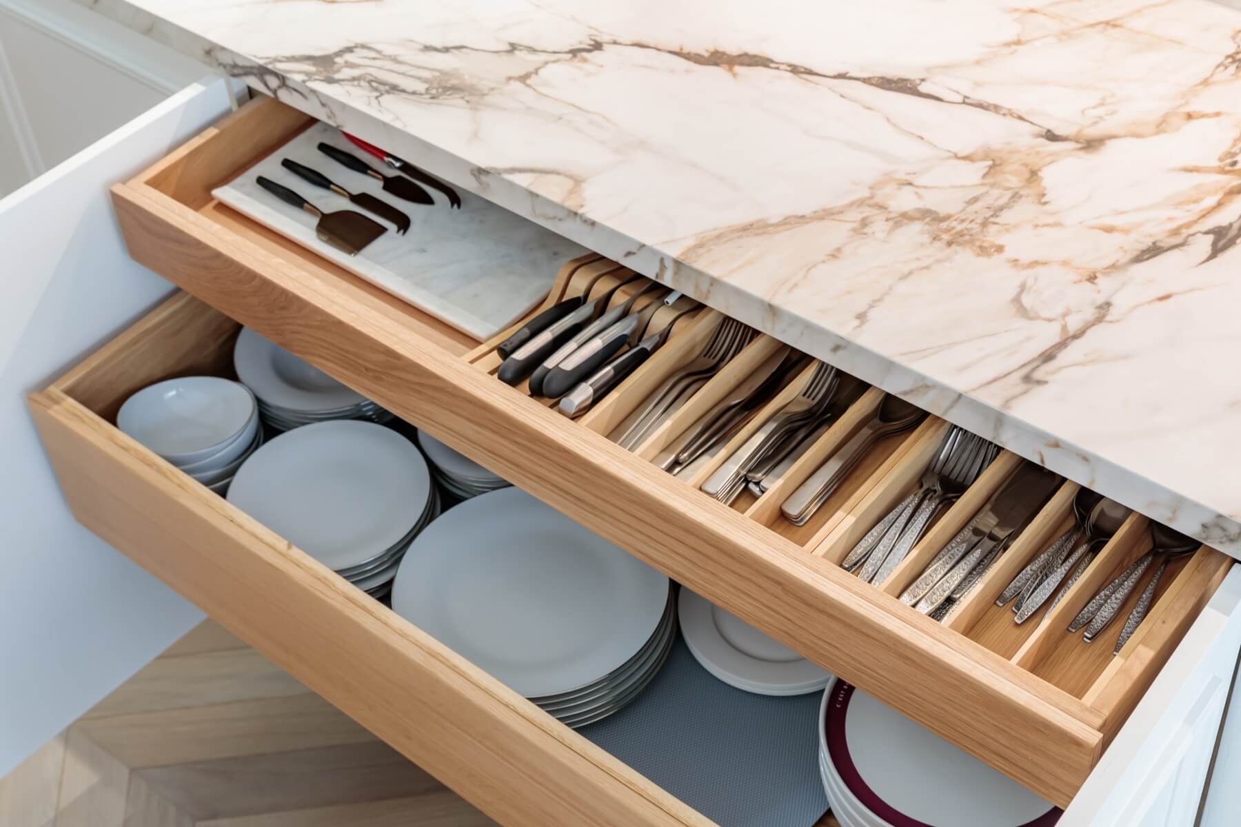 Interior view of a kitchen drawer showcasing neatly organised cutlery and utensils alongside a stack of white plates, set against a marble countertop. An Arch KBB team member is present, contributing to the kitchen design.