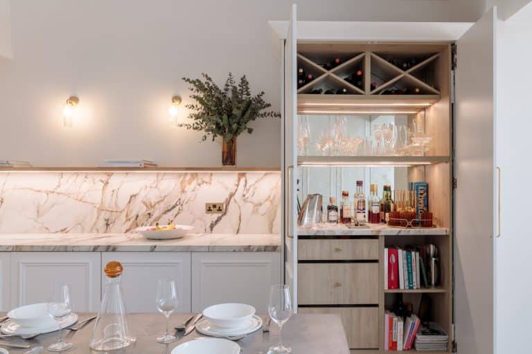 A modern kitchen featuring a marble countertop, elegant lighting, and a stylish cabinet with wine storage and glassware. An Arch KBB team member is present, enhancing the design ambiance.