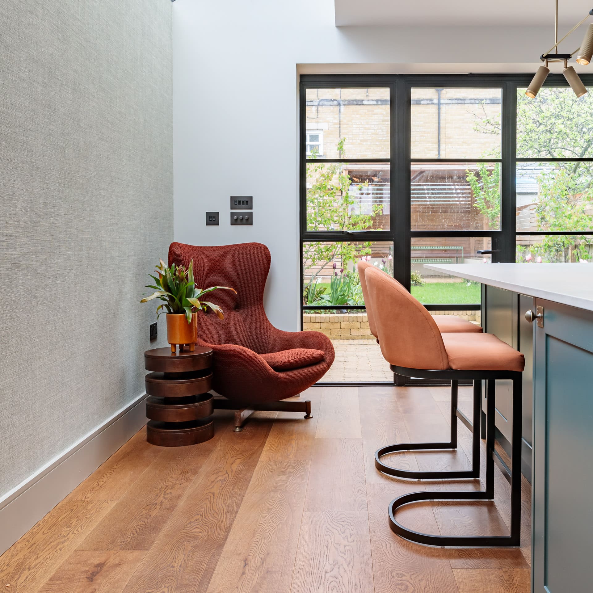 Interior view of a modern kitchen featuring a stylish red accent chair beside a small table with a potted plant, alongside contemporary bar stools. Large windows provide natural light and a view of the garden outside. An Arch KBB team member is present in the space.