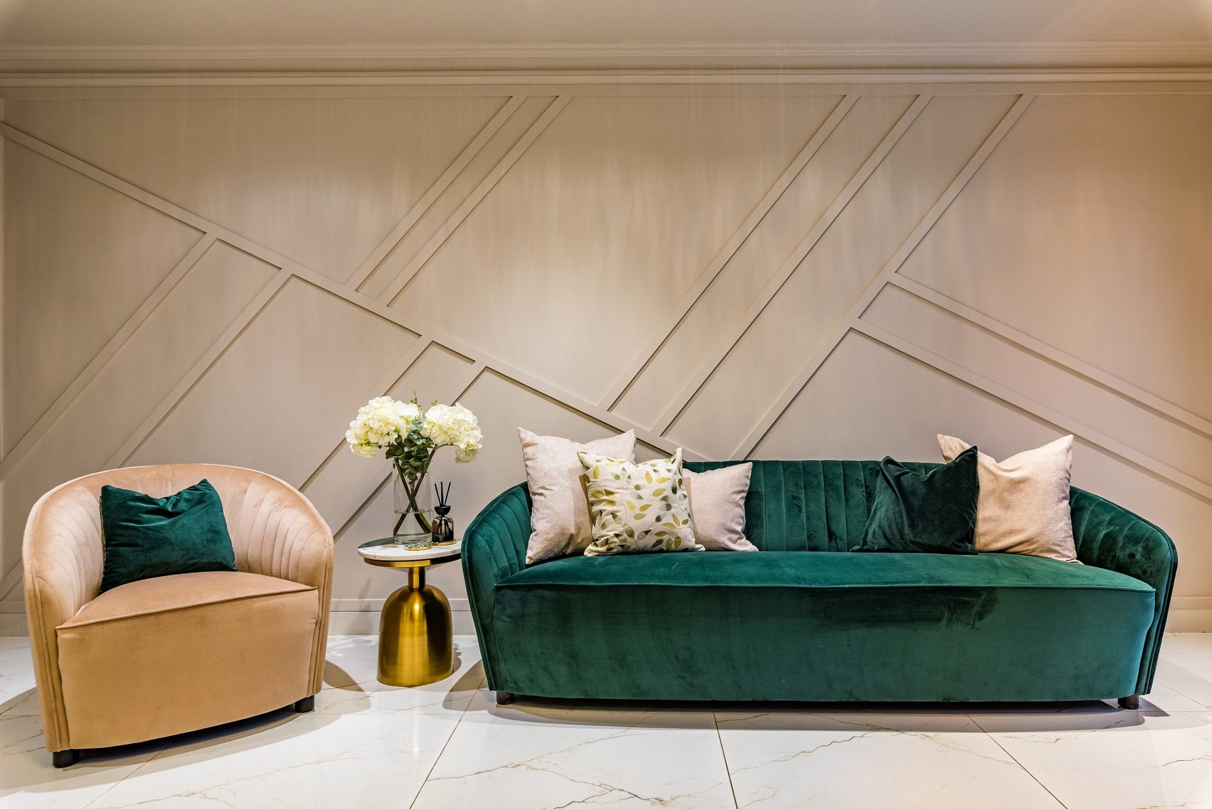 Stylish interior featuring a green velvet sofa and a beige armchair, complemented by a gold side table with a floral arrangement. The backdrop showcases a textured wall design, creating a modern and inviting atmosphere. Arch KBB team member present, enhancing the design aesthetic.