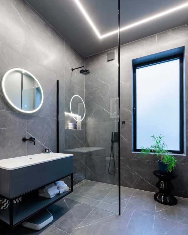 Modern bathroom featuring a walk-in shower with a sleek glass enclosure, marble-effect tiles, a stylish round mirror with integrated lighting, and a minimalist vanity unit. Natural light streams in through a window, complemented by a potted plant for a touch of greenery. An Arch KBB team member is present in the space.