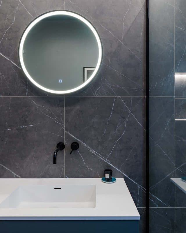 Modern bathroom design featuring a sleek basin, stylish black taps, and a circular illuminated mirror against a dark marble wall. An Arch KBB team member is present, showcasing contemporary interior elements for a sophisticated look.