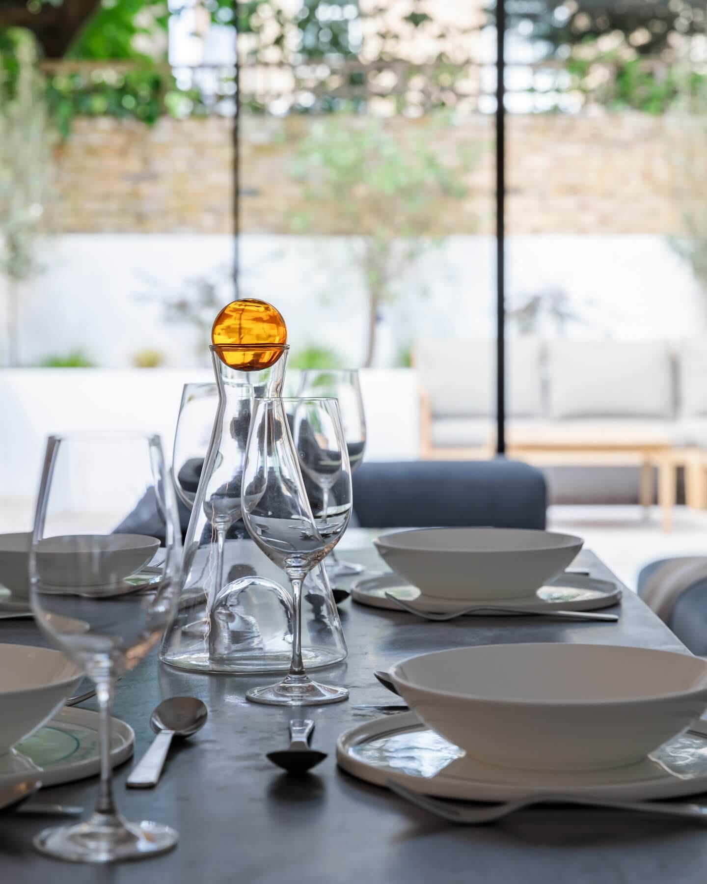 Table setting featuring elegant glassware and minimalist tableware, with an amber decorative piece at the centre. The background showcases a modern outdoor space with seating, enhancing the overall aesthetic. Arch KBB team member present in the setting.