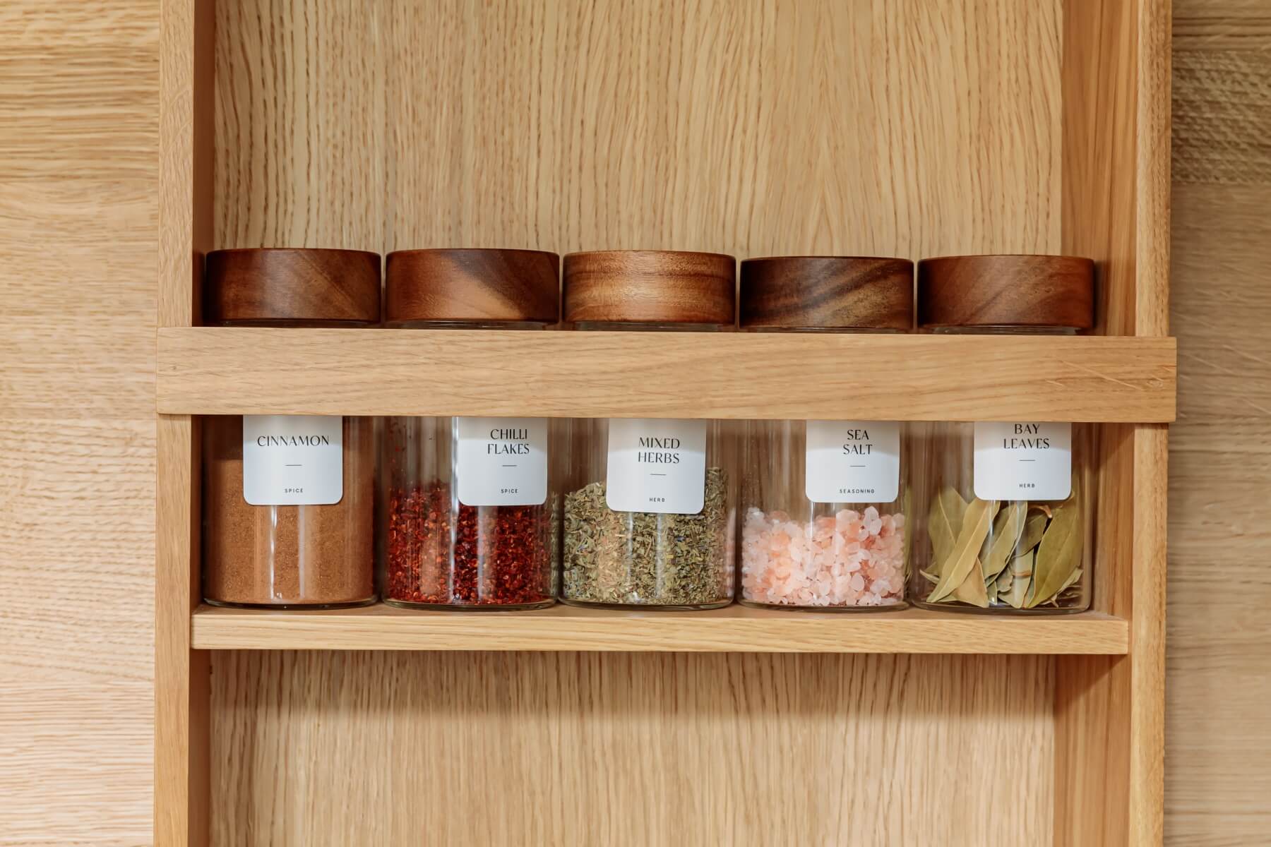Spice jars organized on a wooden shelf, featuring labels for cinnamon, chilli flakes, mixed herbs, sea salt, and bay leaves, showcasing a tidy and functional kitchen storage solution.