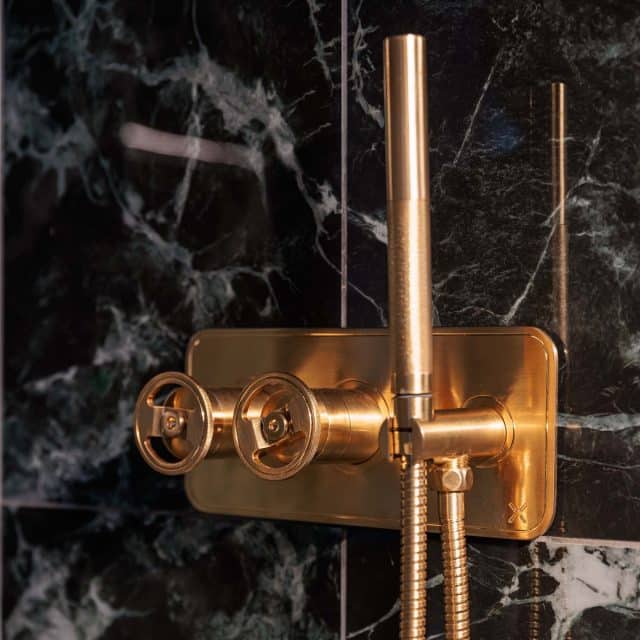 Close-up of a luxurious gold shower control panel mounted on a textured dark marble wall, showcasing two circular knobs and a sleek spout, designed for modern bathroom aesthetics. An Arch KBB team member is present, highlighting the installation process.