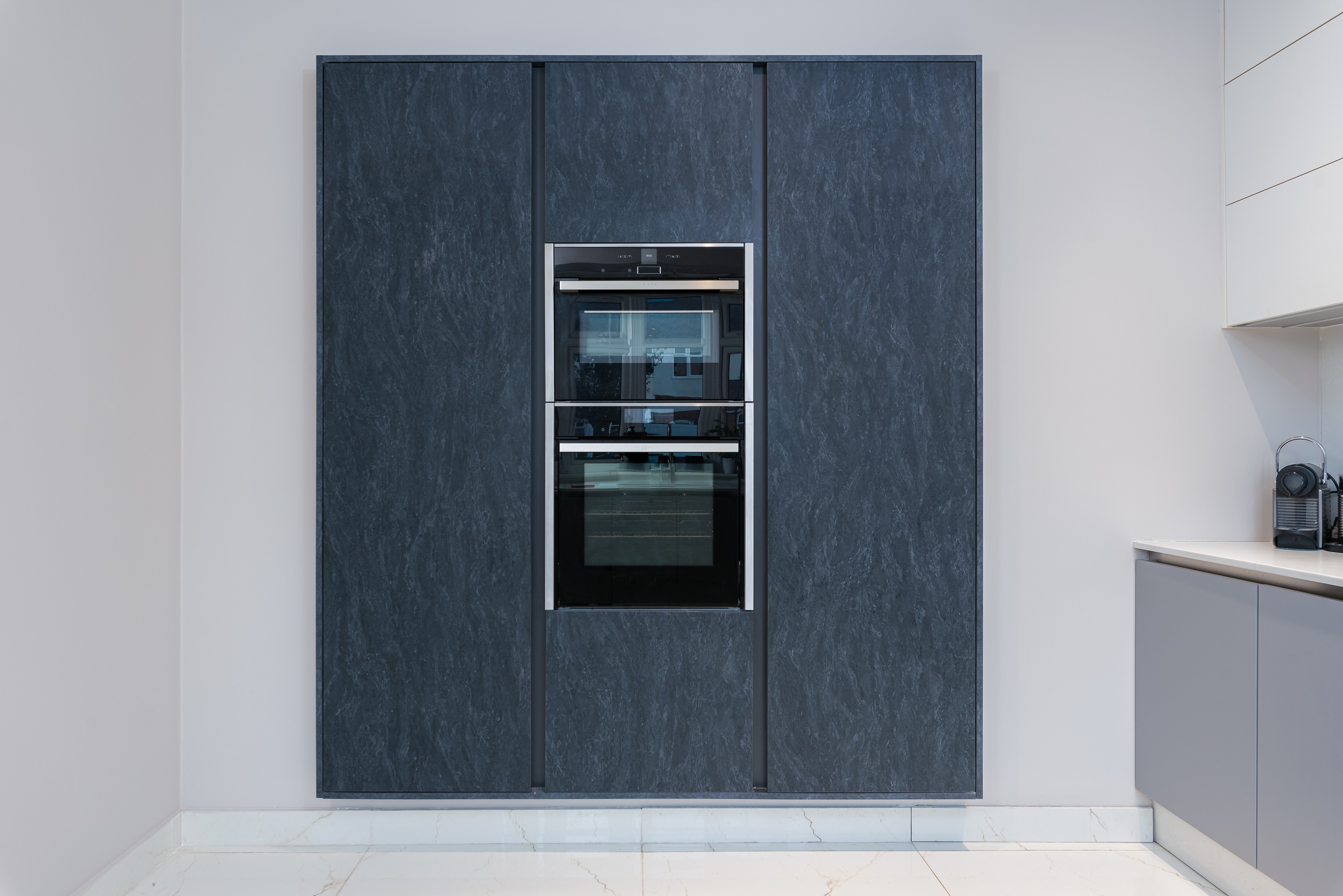A sleek, modern kitchen featuring a dark stone-effect integrated oven unit, surrounded by matching cabinetry. The design showcases a seamless aesthetic, ideal for contemporary interiors. An Arch KBB team member is present, highlighting the kitchen's functionality and style.
