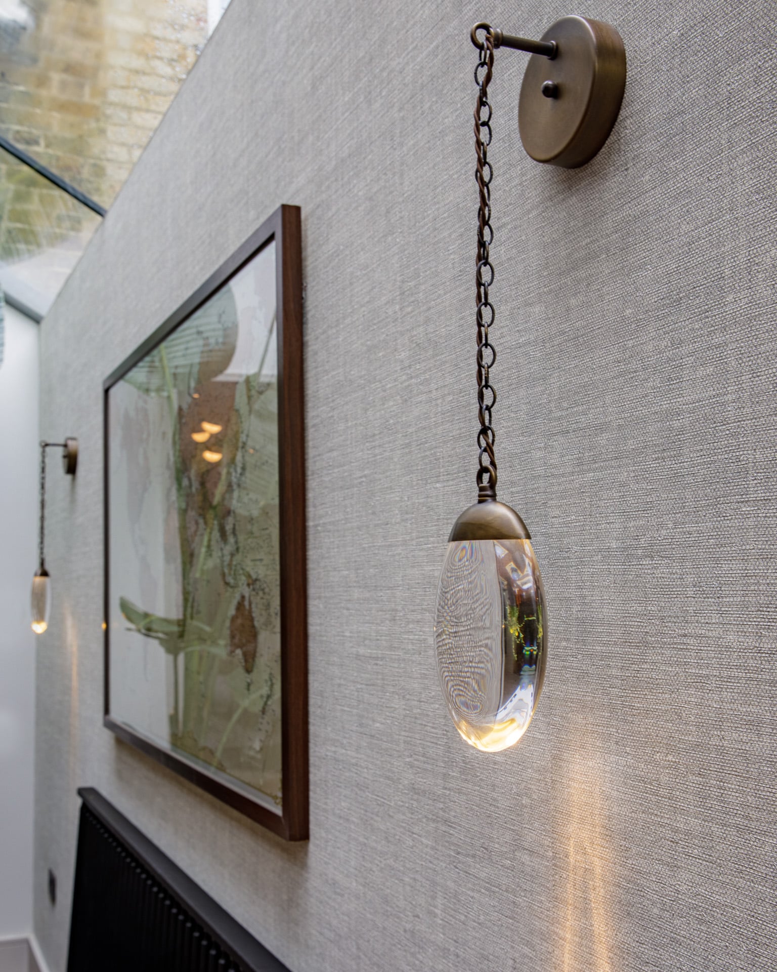 Close-up of a decorative pendant light hanging from a chain against a textured wall, with a framed artwork visible nearby. The light features a clear, oval glass design that reflects ambient light, adding an elegant touch to the interior space.