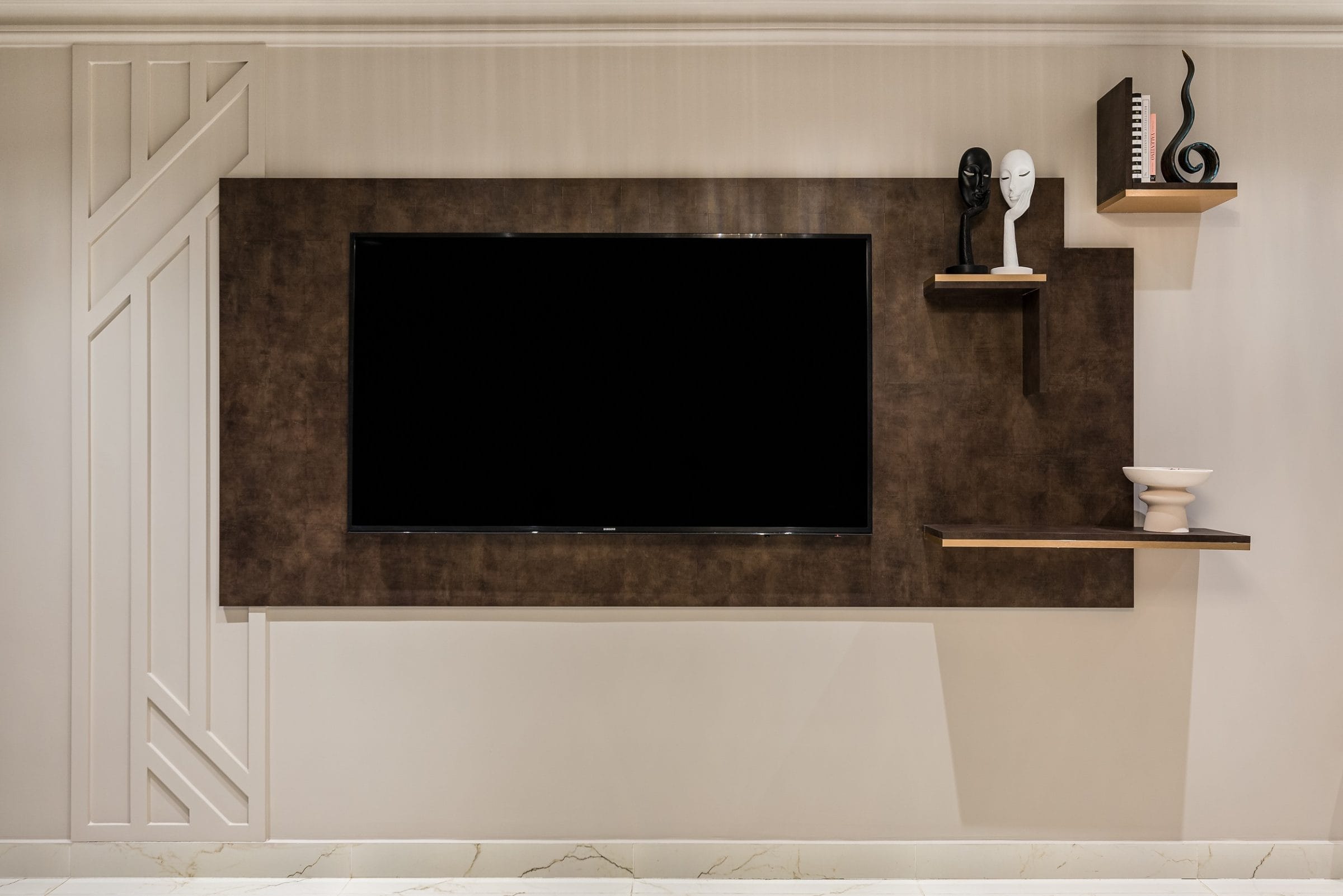 Modern living room with a wall-mounted television and stylish decorative shelves. The backdrop features a sleek brown panel, complemented by minimalist decor items, including sculptures and a small vase. An Arch KBB team member is present, showcasing the design's functionality and aesthetic appeal.