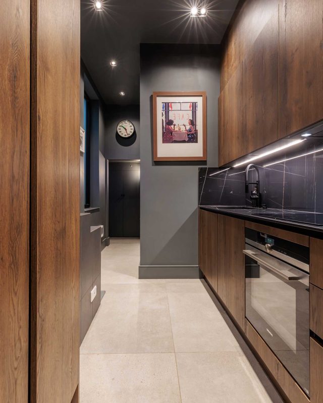 Modern kitchen interior featuring sleek cabinetry, dark tones, and stylish finishes. The space includes a wall clock, framed artwork, and high-quality appliances, showcasing a contemporary design ideal for functional living. An Arch KBB team member is present, contributing to the kitchen's aesthetic appeal.