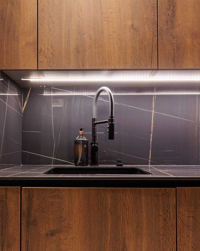 A modern kitchen featuring a sleek black sink with a stylish tap, complemented by a glass bottle and illuminated by under-cabinet lighting. The backdrop consists of elegant dark stone with subtle veining, and warm wooden cabinetry. An Arch KBB team member is present, showcasing contemporary kitchen design elements.