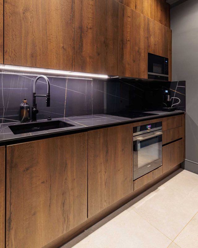 Modern kitchen design featuring sleek wooden cabinetry, a stylish black countertop with gold veining, and integrated appliances. The space includes a contemporary sink and tap, illuminated by under-cabinet lighting, showcasing a blend of functionality and elegance. An Arch KBB team member is present, highlighting the professional service in kitchen renovations.