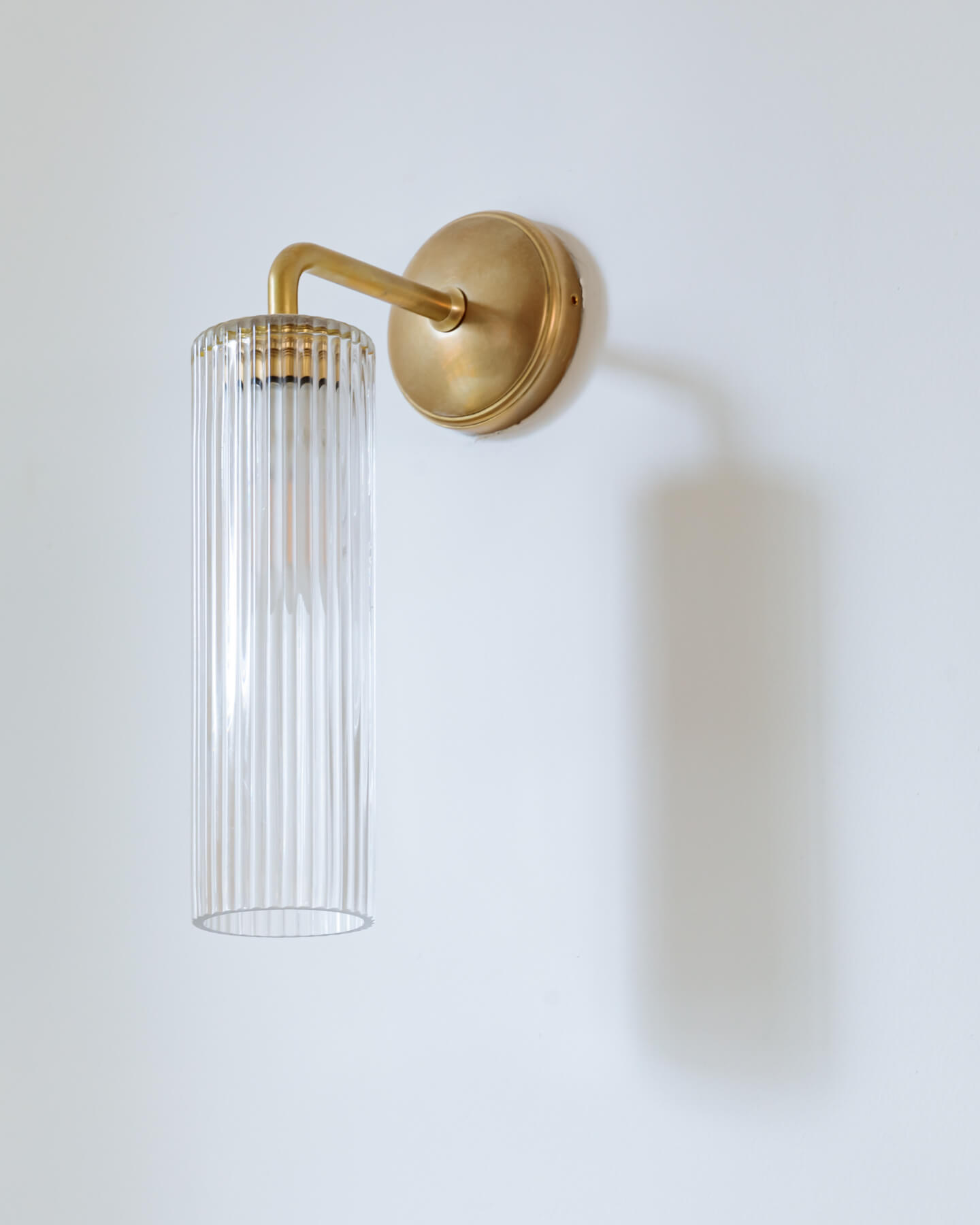 Brass wall light fixture with a clear ribbed glass shade, casting a soft glow against a white wall, designed for modern interior décor.