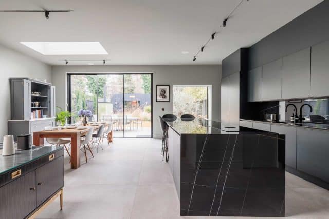 Modern kitchen and dining area featuring sleek cabinetry, an island with a polished black countertop, and large glass doors leading to an outdoor space. An Arch KBB team member is present, enhancing the design experience. Natural light floods the room through a skylight, highlighting the contemporary aesthetic.